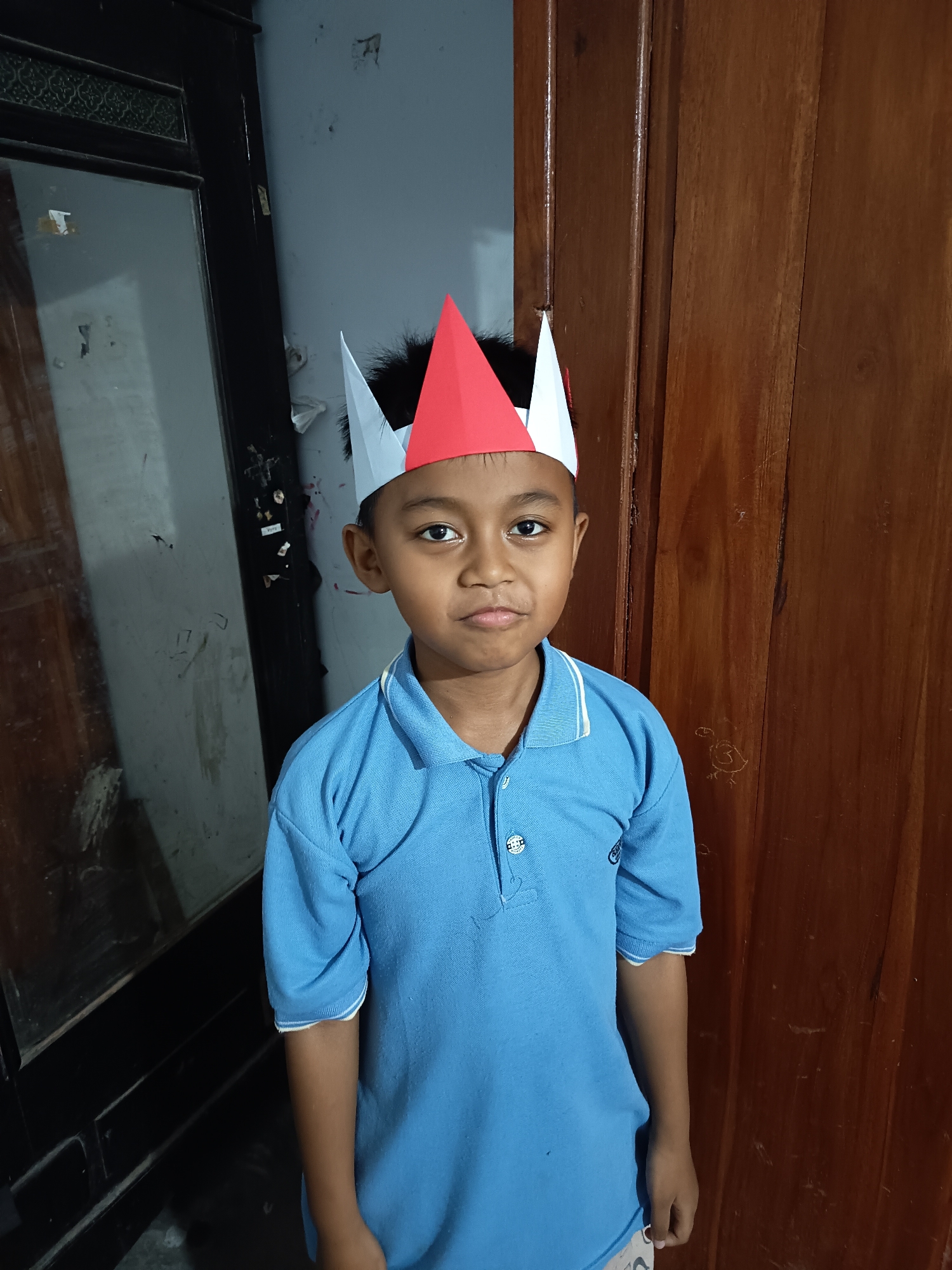 A young boy with dark hair and brown eyes stands in front of a wooden door. He is wearing a blue polo shirt and a red, white, and blue paper crown on his head. The background consists of a dark, wooden door on the right and a dark, reflective surface on the left, possibly a door or a mirror, with a light gray wall behind it. The boy is looking directly at the camera with a neutral expression. The image appears to have been taken indoors, likely in a residential setting in Krampilan, Indonesia, given the presence of traditional Indonesian wooden doors and the appearance of the subject.