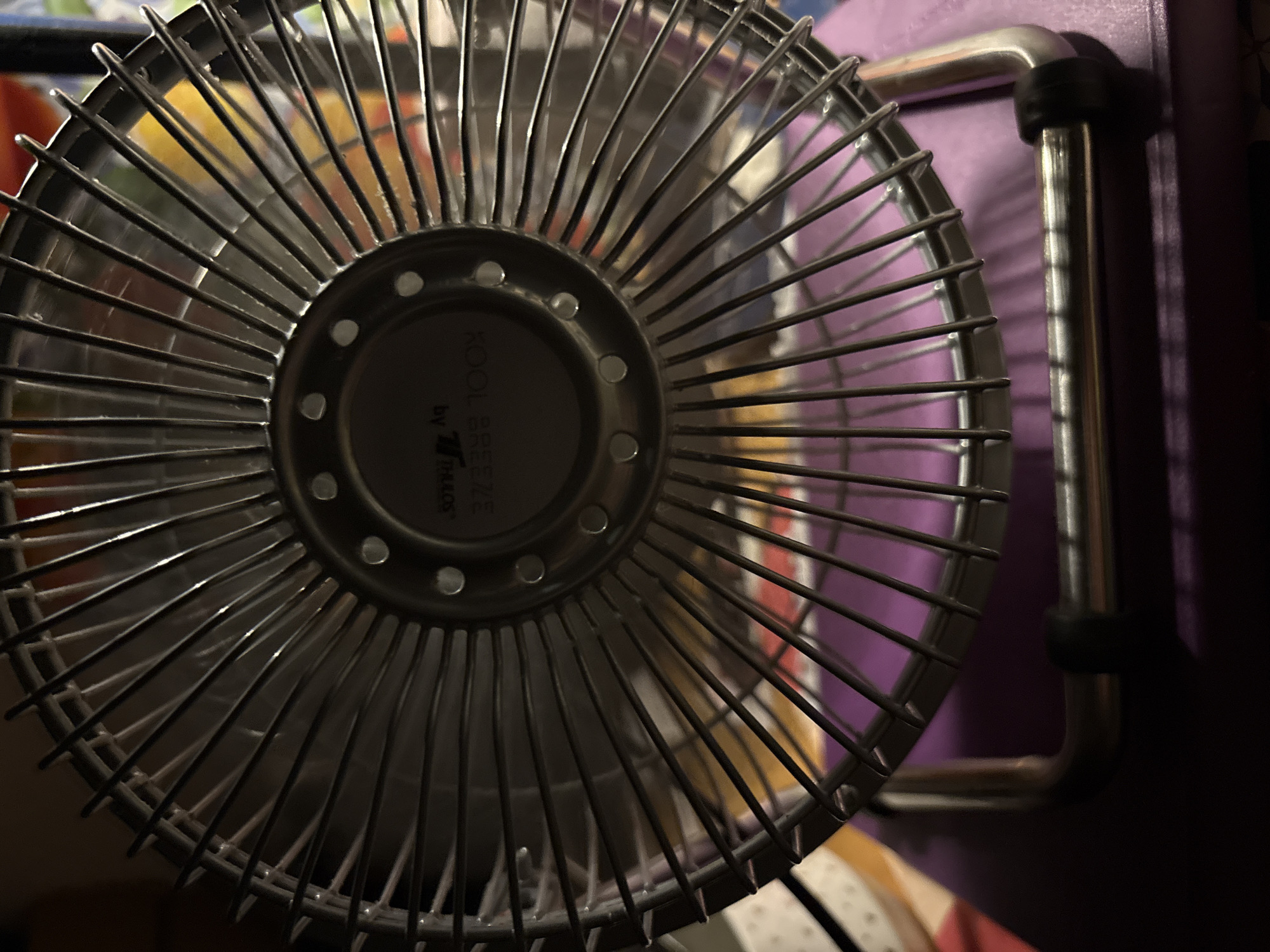 An extreme close-up shot focuses on the metal grille of a small, desk fan. The fan's grille is composed of thin, vertical metal rods arranged radially around a central hub. The central hub appears to be dark gray and has a circular design with some text that is partially visible. There are also small, circular silver accents embedded around the perimeter of the hub.
The fan is positioned in a way that the viewer sees it from slightly above and to the side. The background is blurred and out of focus, but hints of color, including purple and yellow, can be discerned. The overall lighting is dim, with highlights reflecting off the metal surfaces of the fan grille, creating a slightly glossy and reflective appearance. The fan is resting on a surface that appears to be purple. The bottom of the fan's metal stand is visible in the foreground, with a black rubber stopper on each side. The image has a shallow depth of field, with the fan grille being the sharpest element.
