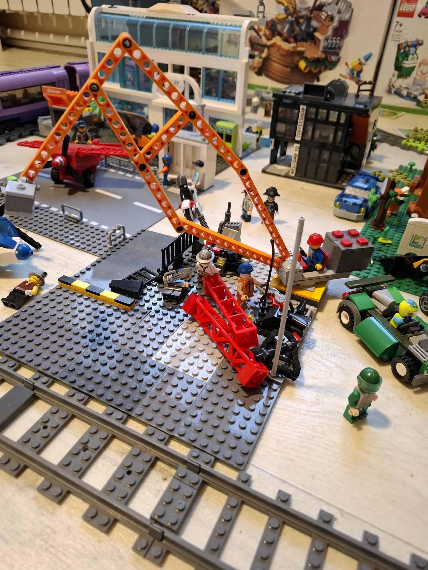This is a high-angle, medium shot of a Lego construction scene. In the foreground, there are Lego train tracks. Behind the tracks, a large grey Lego baseplate is laid out, with a variety of Lego elements scattered across it. These include minifigures in various poses and clothing, some appearing to be working on a construction project. There is a red Lego crane with orange supports, and a red Lego ramp. Other Lego vehicles, such as a green race car with a yellow-headed driver and a blue car, are present. In the background, there is a multi-story Lego building with a glass facade and a sign that reads "COFFEESHOP." Another Lego building, white with blue windows, is to the left, and a purple Lego train is visible behind it. The scene is set on a light-colored, wooden surface, resembling a floor or table. There is no indication of the city of Tyumen, Russia.