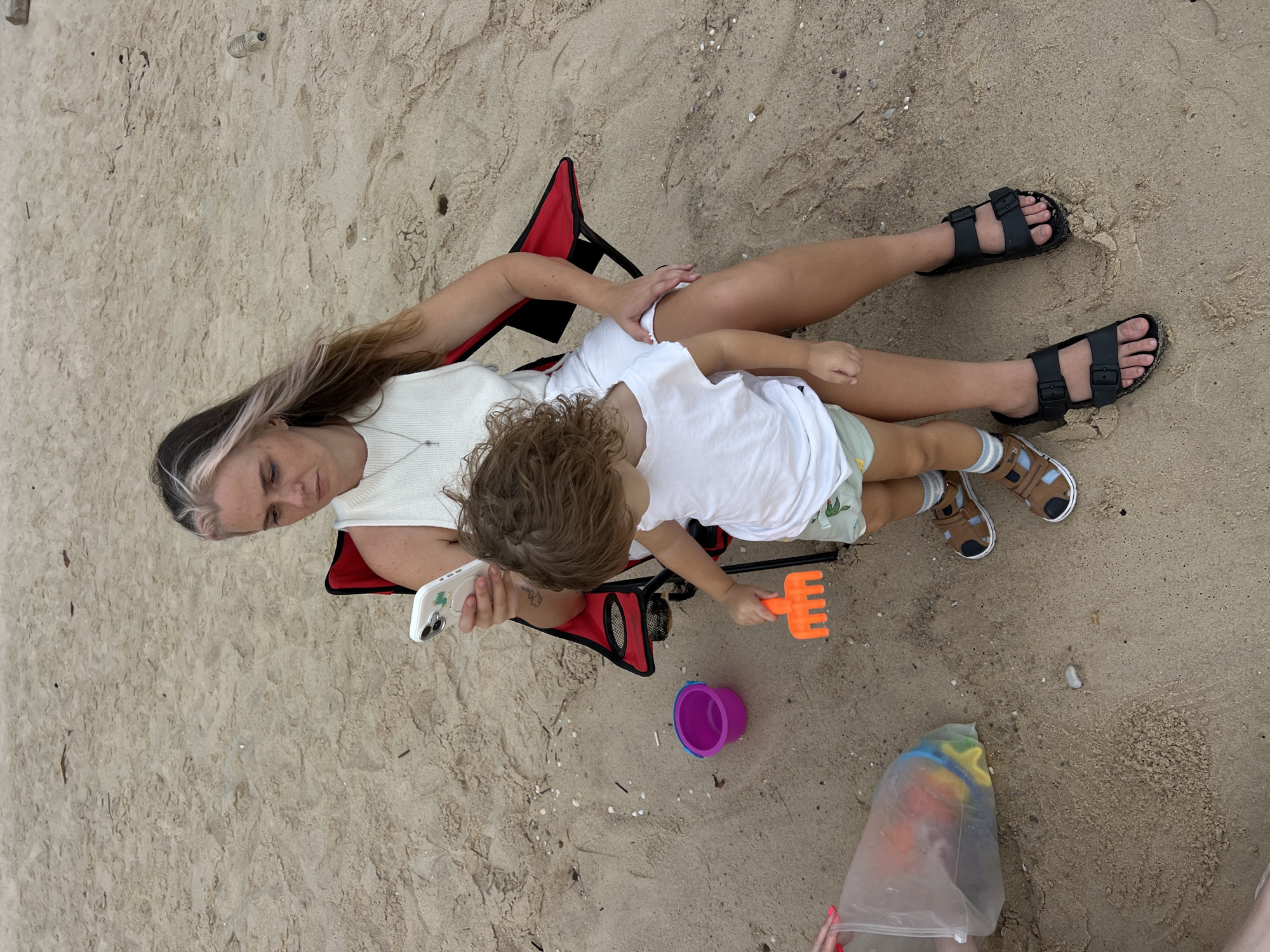 An overhead shot shows a woman and a young child sitting on a beach. The woman is sitting in a red folding chair, and the child is sitting on her lap. The woman is wearing a white top and black sandals. The child is wearing a white t-shirt and shorts. The child is holding an orange plastic rake. In front of the child is a purple bucket. To the right of the child, a plastic bag filled with colorful toys is visible on the sand. The woman is holding a white smartphone in her left hand. The scene is set on a sandy beach with some shells and debris scattered across the sand. The lighting suggests it is daytime.