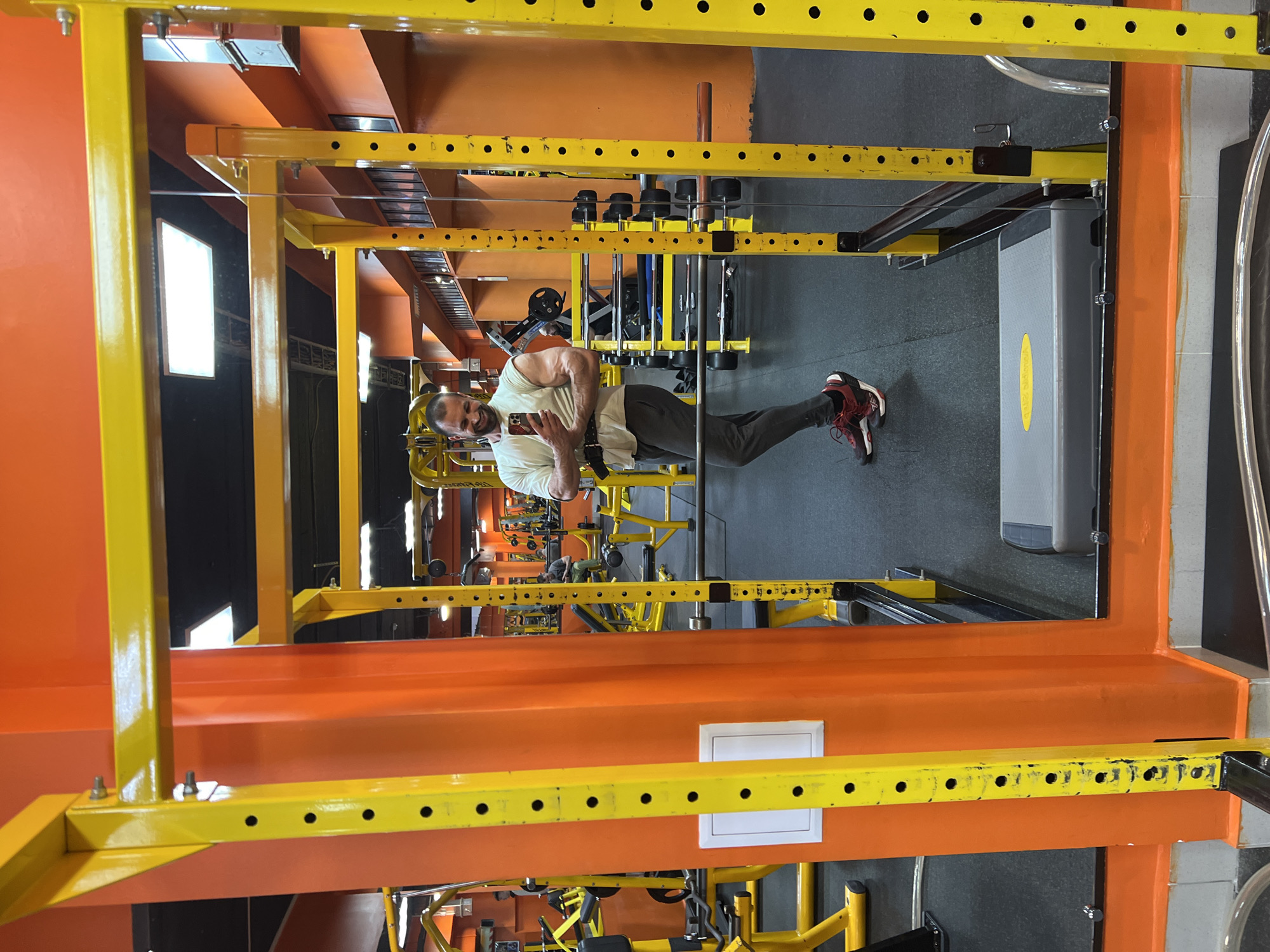 The image shows a man in a gym in Yoshkar-Ola, Russia. The gym has orange and yellow equipment, including a power rack and weights. The man is holding a phone, possibly taking a selfie. He is wearing athletic clothing and shoes. The gym has a gray floor and some natural light from windows.