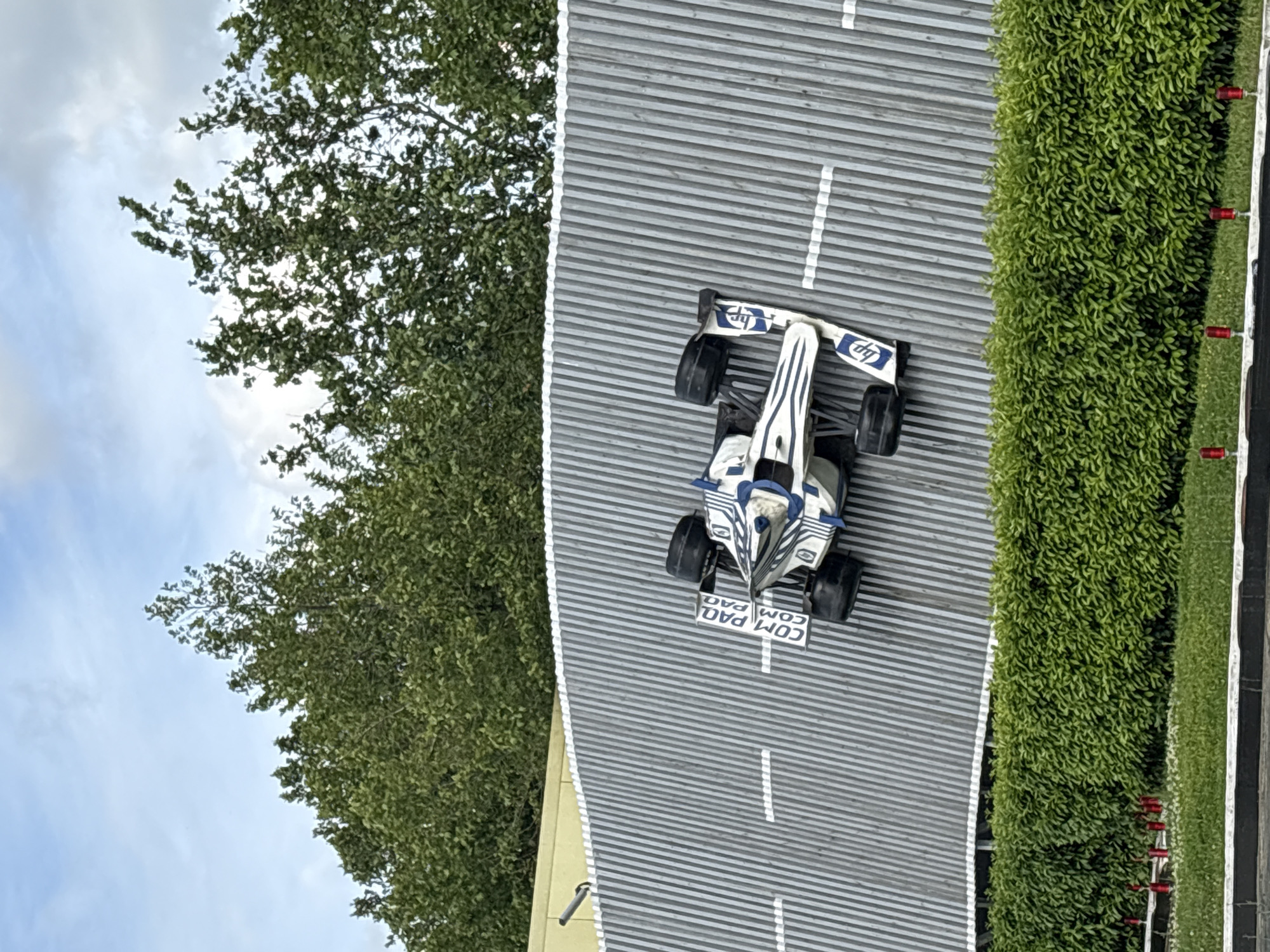 An aerial, oblique shot reveals a miniature model racing track in Anapa, Russia, bordered by meticulously manicured shrubs on the right and leafy trees on the left, suggesting a park or public space. A white and blue Formula One-style race car, branded with "COMPAQ" and "hp" logos, is centrally positioned on the grey, striped roadway. The road features white lane markings. A portion of a partly cloudy sky is visible in the upper left corner.