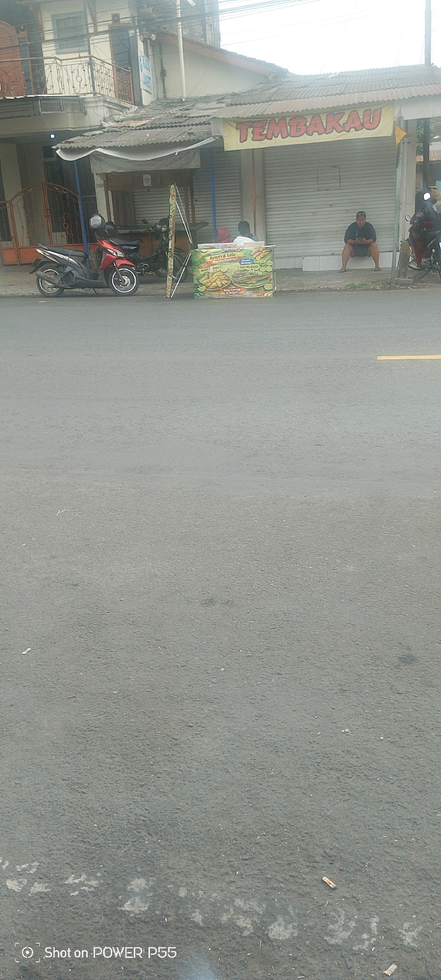 In the city of Unknown, Unknown, a street scene unfolds with a focus on a small roadside food stall. The stall, adorned with a banner displaying "TEMBAKAU," appears to be selling fried chicken and catfish, as indicated by the imagery on its display. Two individuals are visible near the stall: one seated on a low wall, engrossed in their phone, and another partially obscured by the stall's structure. A red motorcycle is parked to the left of the stall, and another motorcycle is visible to the right, suggesting a mode of transportation for patrons or vendors. The street itself is paved, with the right side of the road marked by a yellow line. The environment depicts a typical urban setting with buildings lining the street, some showing signs of age and wear. The sky is overcast, contributing to the muted lighting of the scene. The overall impression is of a snapshot of daily life in a busy commercial area.