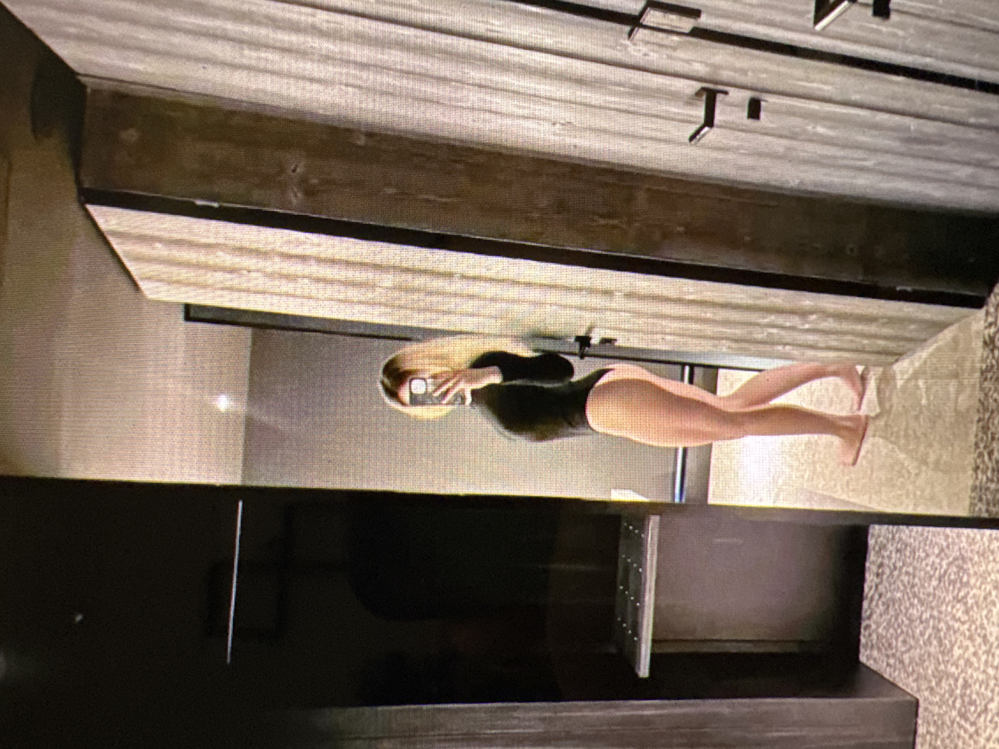 The image shows a woman taking a selfie in a mirror. She is wearing a black long-sleeved bodysuit and appears to be in a bathroom. The room has modern finishes, including what looks like wood-paneled walls and a tiled floor. There are multiple doors visible in the reflection. The woman is positioned in the center of the frame, with her back partially turned to the camera as she holds a phone to take the photo. The lighting suggests an indoor setting, possibly a hotel room or a private residence. The image is pixelated, indicating it may be a screenshot or a low-resolution photograph. The city of Castelo Branco, Portugal, is not depicted in the image, nor is there any information available to suggest this location.