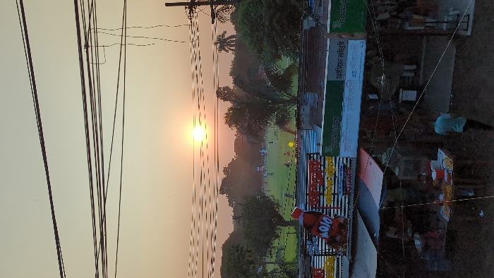 The image captures an urban landscape in Savar, Bangladesh, during either sunrise or sunset, indicated by a low, bright sun in a hazy, warm-toned sky. The foreground features a street-level commercial area comprising multiple stalls or small shops under corrugated metal roofs and fabric awnings. A food stall displays various prepared snacks on a table. One individual in a light blue shirt is observed walking away from the stall area on the right, with other indistinct figures present. Overhead signage includes a large "MOJO" beverage advertisement and green banners with the text "GOOD LUCK" in English, accompanied by Bengali script that partially translates to "Good Luck Library" and "Official Market and Stationery." Above this commercial layer, a expansive green field, appearing to be a park or sports ground, is visible. Multiple small groups of people are scattered across the field, some engaged in recreational activities resembling cricket or football, with a yellow spherical object (possibly a ball) on the grass. The field is bordered by numerous trees and foliage in the mid-ground and background. The upper section of the image is characterized by a dense network of dark electrical and utility wires, with a utility pole on the far right.