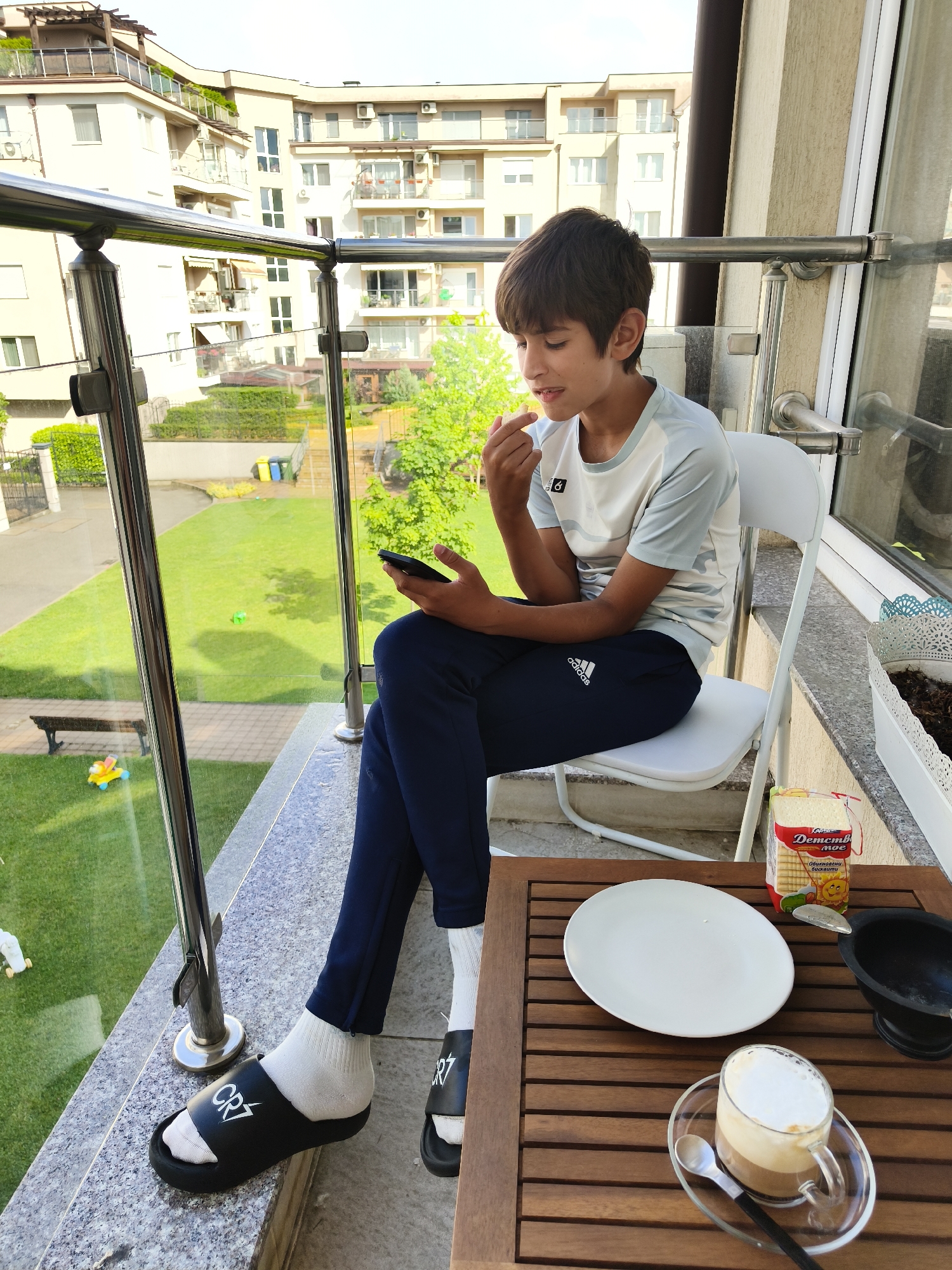 A young boy with short dark hair is seated on a white folding chair on a modern balcony during the daytime. He is wearing a light-colored t-shirt with a small black logo (number 6 visible), dark blue Adidas sweatpants, white socks, and black CR7 branded slides. He holds a black smartphone in his left hand and his right hand is near his mouth, suggesting he might be eating or picking at something.
The balcony features sleek metal and glass railings and a grey tiled floor. To the boy's right, a slatted wooden table holds a white oval plate, a clear glass mug containing a layered coffee drink (likely a latte or cappuccino) on a matching saucer with a spoon, a small black bowl or mortar, and a packet of biscuits. The biscuit packaging is red and white, displaying Cyrillic text that reads "Детство мое" (My Childhood) and "Обикновени бисквити" (Ordinary Biscuits), along with a smiling sun logo.
Beyond the balcony, a well-maintained green lawn is visible, featuring a yellow and blue child's toy and a green plastic object. Further back, a paved area with a wooden bench is seen. In the background, several modern multi-story residential buildings with balconies, pergolas, and air conditioning units are prominent, surrounded by trees and lush greenery. The scene depicts a calm, relaxed moment in an urban residential setting, likely in Sofia, Bulgaria, given the visible text and architectural style.