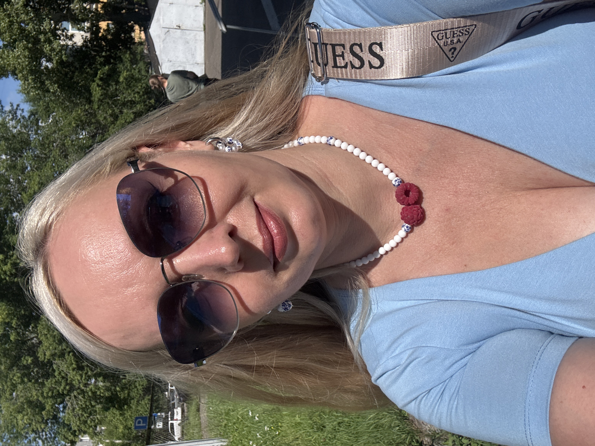 The image is a selfie of a blonde woman with sunglasses and a light blue top. She is wearing a necklace with white beads and two red raspberry-shaped pendants, as well as hoop earrings with a blue and white pattern. A beige strap with the "GUESS" logo is visible across her shoulder. The background is an outdoor scene with green trees, grass, a fence with a parking sign, and a white building. In the distance, another person in a green shirt can be seen. The lighting suggests it is daytime and sunny. There is no visual information in the image that can identify the location as Komsomolsk-on-Amur, Russia.