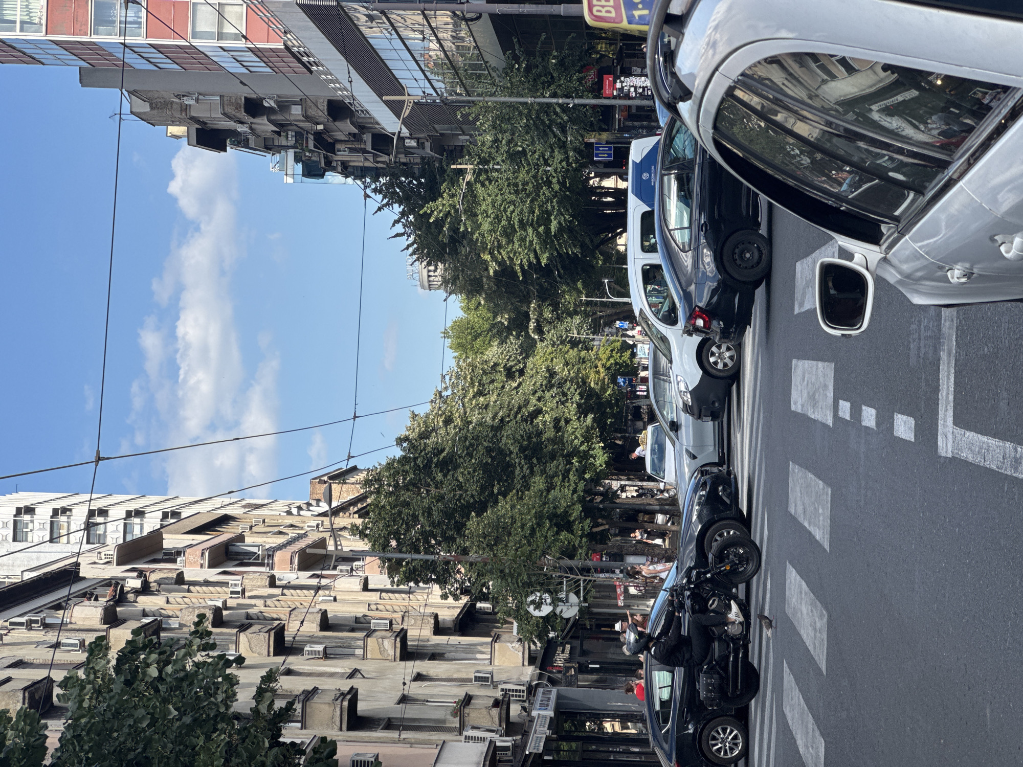 The image captures a busy street scene in Belgrade, Serbia, likely near the intersection of Trg Nikole Pašića and Boulevard of King Alexander. The perspective is from a vehicle in the foreground, looking through a zebra crossing at a traffic jam. On the left, older apartment buildings with multiple balconies and visible air conditioning units line the street. On the right, a modern building with a red facade and glass windows stands out.
Several vehicles are present: a motorcycle with a rider wearing a black helmet and jacket is in the left lane, a black sedan is behind it, and other cars and a white van are visible further down the street, contributing to the traffic congestion. People are seen on the sidewalk and crossing the street. A pigeon is walking on the zebra crossing in front of the motorcycle. Overhead, power lines crisscross the sky, and lush green trees provide shade along the street. The sky is blue with scattered white clouds. A yellow taxi is also visible among the vehicles.