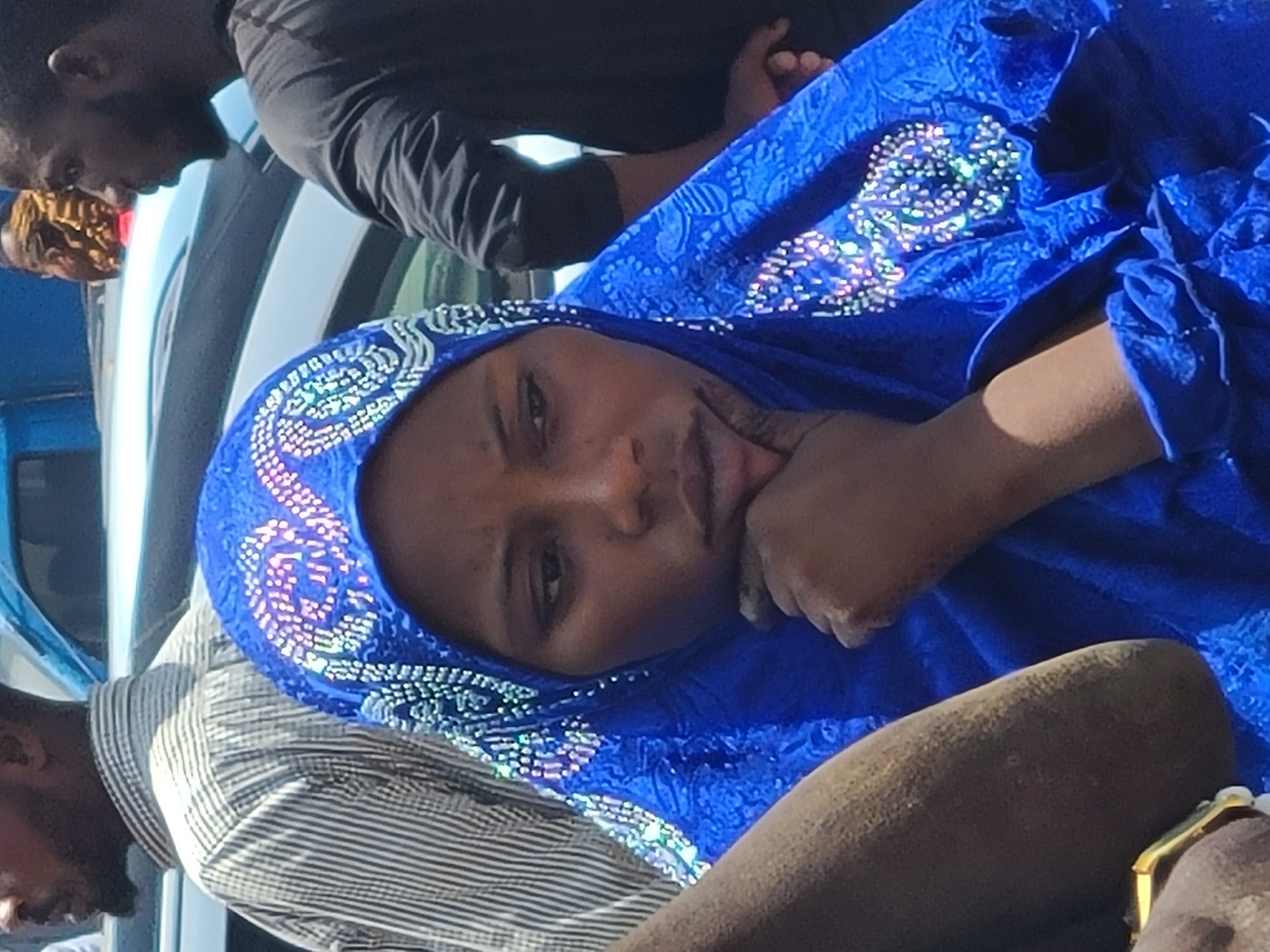 In the city of Jos, Nigeria, a young woman wearing a blue hijab with sparkly embellishments rests her chin on her hand, her gaze directed downwards, suggesting contemplation or sadness. The hijab is intricately decorated with a pattern of white beads and reflective elements, creating a shimmering effect. Her skin tone is dark, and her features are full. To her left, another person's arm, also dark-skinned, is visible, with a gold-toned bracelet on their wrist. Behind her, a man in a striped shirt is partially visible, and to the far right, another man in a dark t-shirt stands with his mouth open, as if speaking or reacting to something. The background reveals the glint of car lights, indicating an outdoor setting likely near a street or parking area. The overall scene is a candid moment captured in a public space, with the focus on the expressive face of the woman in the hijab.