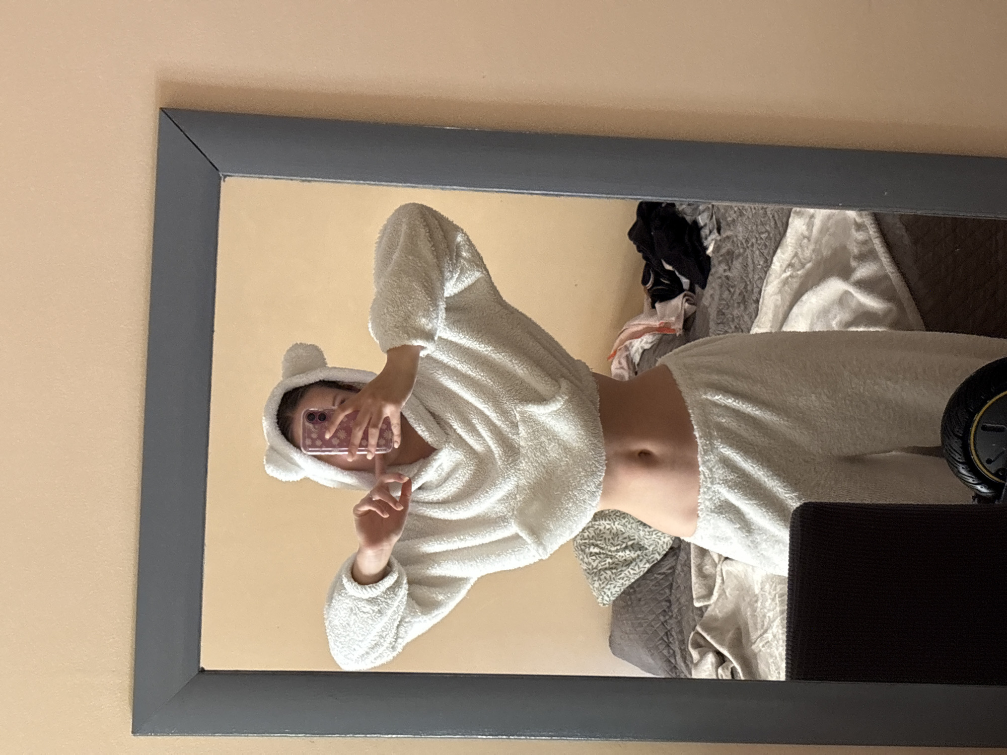 This image captures a mirror selfie taken indoors in Bečići, Montenegro. The primary subject is a person, whose face is obscured by the smartphone they are holding to take the picture. They are dressed in a cozy, white, two-piece loungewear set made of a fluffy, towel-like material. The set includes a cropped hoodie with small, rounded "bear" ears on the hood, revealing their midriff, and matching long pants. One hand is holding the phone, while the other hand is positioned as if making a gesture.
The mirror, framed in dark gray, reflects an indoor setting, likely a bedroom or private living space. The walls visible in the reflection are a light, warm beige or peach color. Behind the person, a bed is partially visible, covered with rumpled blankets in shades of gray and white, along with some piled clothes (dark and light fabrics), indicating a casual, lived-in environment. To the right, a dark object with a yellow circular detail, possibly a wheel of a scooter or a similar piece of equipment, is visible. The lighting is soft and indoor, and no specific time of day can be determined. There is no visible text in the image.