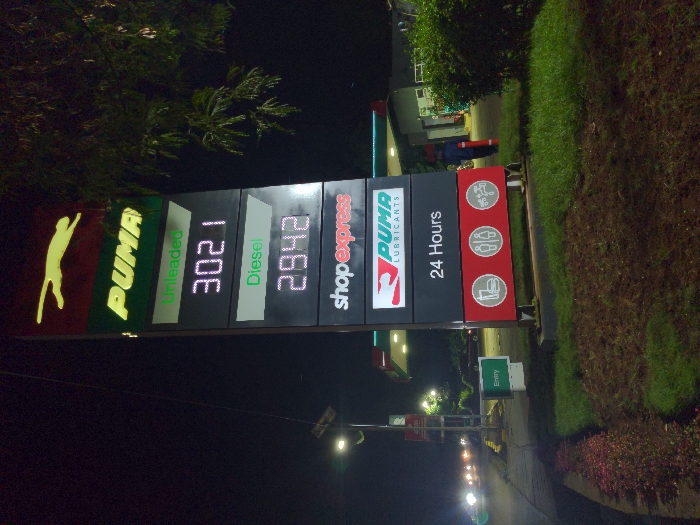The image shows a Puma gas station sign at night. The sign displays the price of unleaded gas at 302.1 and the price of diesel at 294.2. The sign also indicates that the gas station is open 24 hours and has a shop express, and facilities for restrooms and car wash. A single individual can be seen walking on the asphalt of the gas station, away from the sign. The location is a gas station in Lindi Street, Tanzania, with coordinates -3.352 latitude and 37.338 longitude. The gas station is located in a well-lit area with lush green grass and trees. The ground appears to be asphalt, indicating a paved parking lot.