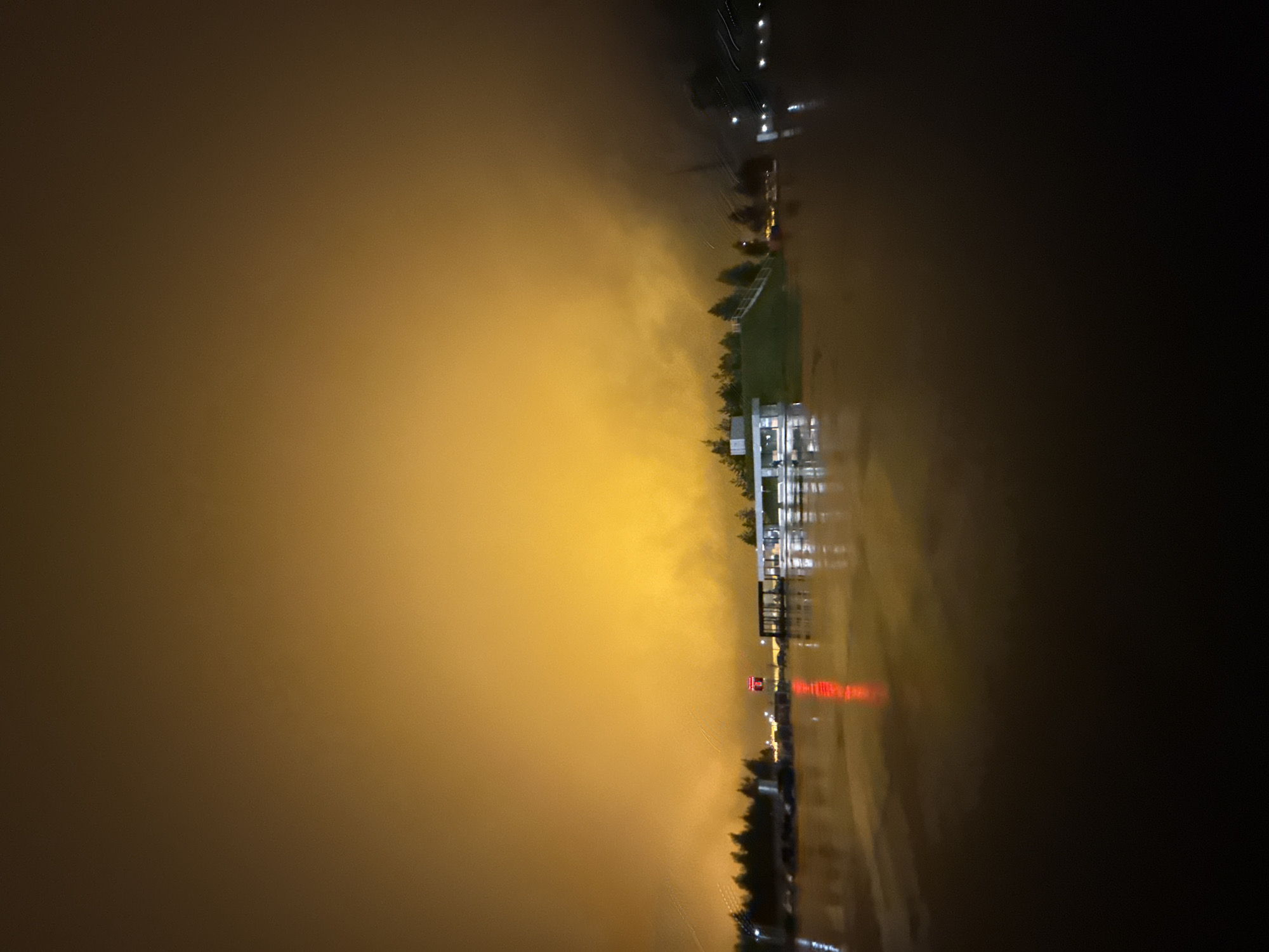 The image depicts a nighttime scene in Sarapul, Russia, characterized by an intensely illuminated, orange-yellow sky suggesting heavy light pollution reflected by a low cloud cover or dense fog. The foreground is dominated by a dark body of water, likely a river or a flooded area, which exhibits reflections of various distant lights.
Along the horizon, a line of dark silhouettes of conifer trees is visible on both the left and right sides. In the center-left, a modern, light-colored building with large windows is situated on a low, grassy mound, possibly an embankment, and its lights are reflected in the water. Further to the right, a prominent vertical red light or sign emits a strong, elongated reflection in the water. To the right of this red light, another structure with multiple internal lights and a more expansive, open design is visible, also casting reflections. Additional faint white and yellow lights are scattered along the distant shoreline, some appearing as streaks due to motion or long exposure. The atmosphere is obscured by the glowing haze, particularly on the right side of the frame where structures become less distinct. No individuals or specific actions are discernible within the frame.