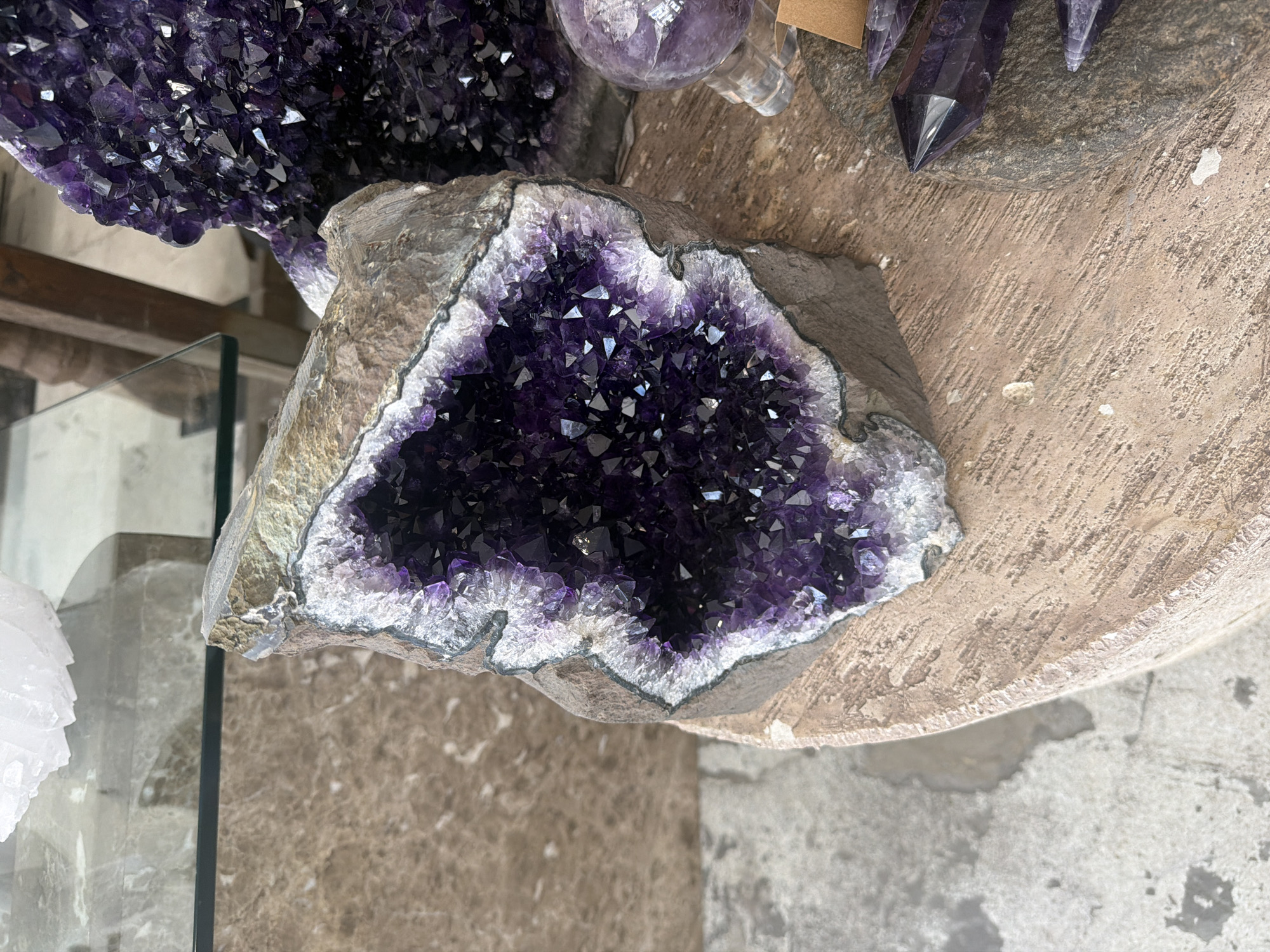 This image shows an amethyst geode with a dark purple crystal interior. The geode is set on what appears to be a textured, light-brown, round surface. In the background, there are more amethyst crystals, including a cluster and pointed crystals. A glass pane and a marble or textured stone surface are also visible in the background. This photograph might be taken in a shop or store selling crystals, possibly in Los Angeles, United States of America. The exact location cannot be determined based on the given image.