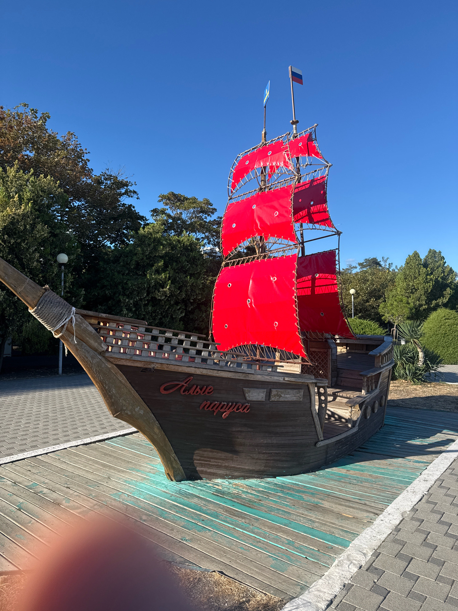 This is a medium-shot, eye-level, outdoor, daytime, vertical, candid photograph of a wooden pirate ship replica with bright red sails, situated on a wooden platform painted with turquoise streaks. The ship has the Russian inscription "Алые паруса" (Alye Parusa), meaning "Scarlet Sails," painted in red cursive on its side. The ship is positioned in the city of Anapa, Russia, likely near the coast, given the presence of palm trees and lush green bushes and trees in the background. The sky is a clear, bright blue, indicating fair weather. The ship appears to be a decorative or playground element rather than a functional vessel, as it is grounded on a wooden deck that merges with a paved walkway. There are no people visible in the frame. The overall scene suggests a tourist attraction or a recreational area.