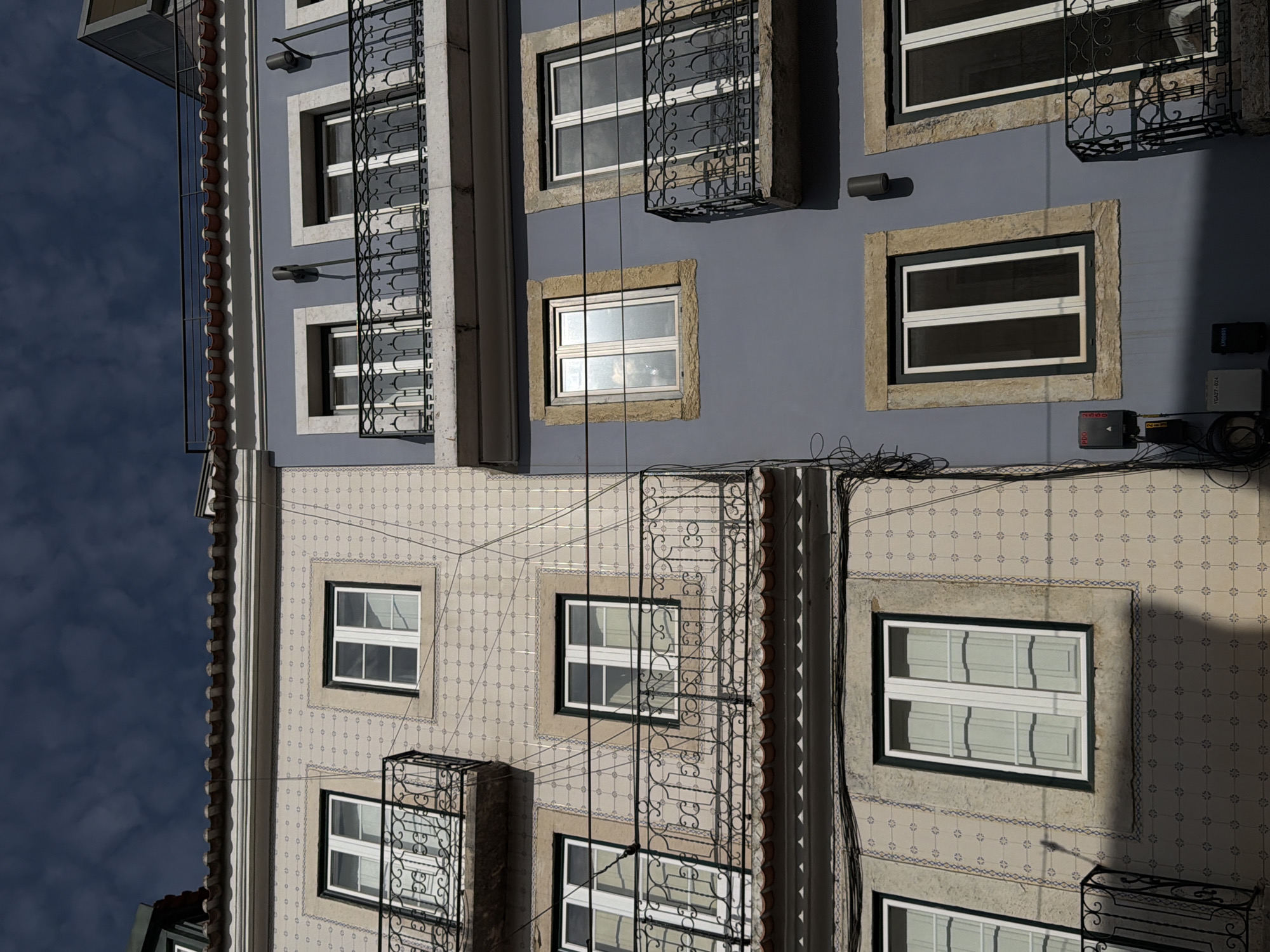The image displays the facades of two adjacent buildings in Lisbon, Portugal, under a cloudy sky. The building on the left features a light-colored tiled exterior adorned with a repeating pattern of small blue squares and intersecting lines. It has several windows, some with white frames and panes, and a decorative black wrought-iron balcony with intricate scrollwork. The building on the right has a smooth, pale blue painted facade. This building also has windows, framed with light-colored stone, and a similar black wrought-iron balcony. Electrical wires and junction boxes are visible along the facades and running between the buildings. The architectural style suggests an older urban setting in Lisbon, characterized by decorative tilework and wrought-iron balconies.