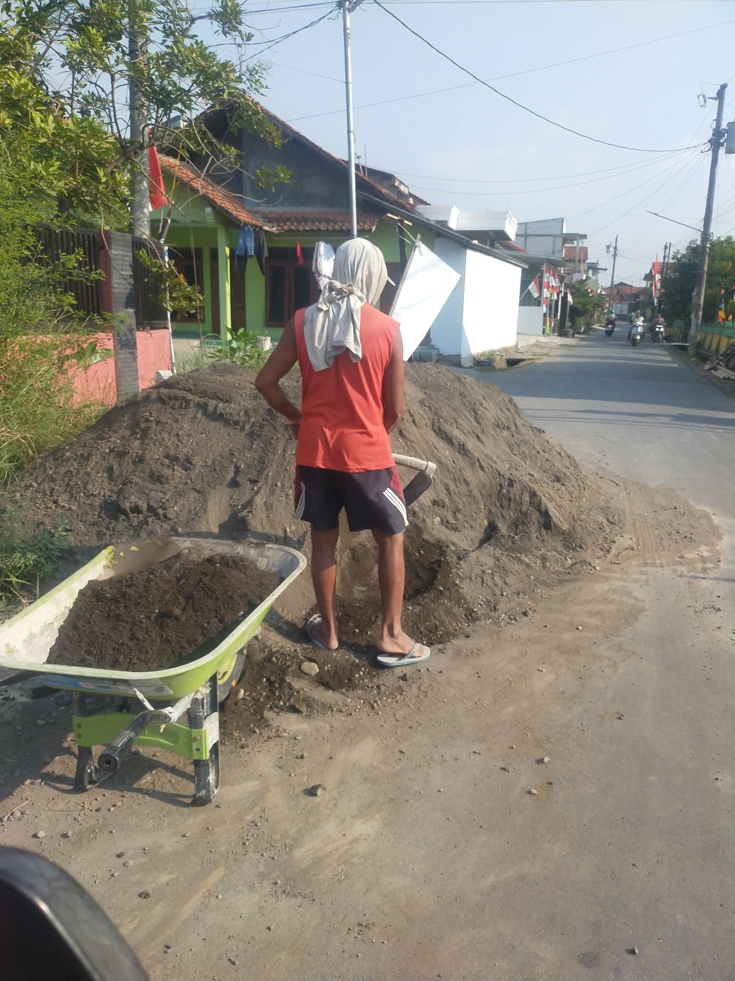 A person wearing a red tank top, dark blue shorts with white stripes, and a white head covering stands on a dirt road in Sumingkir, Indonesia. The person is next to a large pile of dirt and is holding a shovel. In the foreground, a green wheelbarrow filled with dirt is positioned to the left of the person. The background shows houses with red tile roofs, a white building, and trees. A street with several motorcycles is visible in the distance. The scene appears to be a construction or landscaping site.