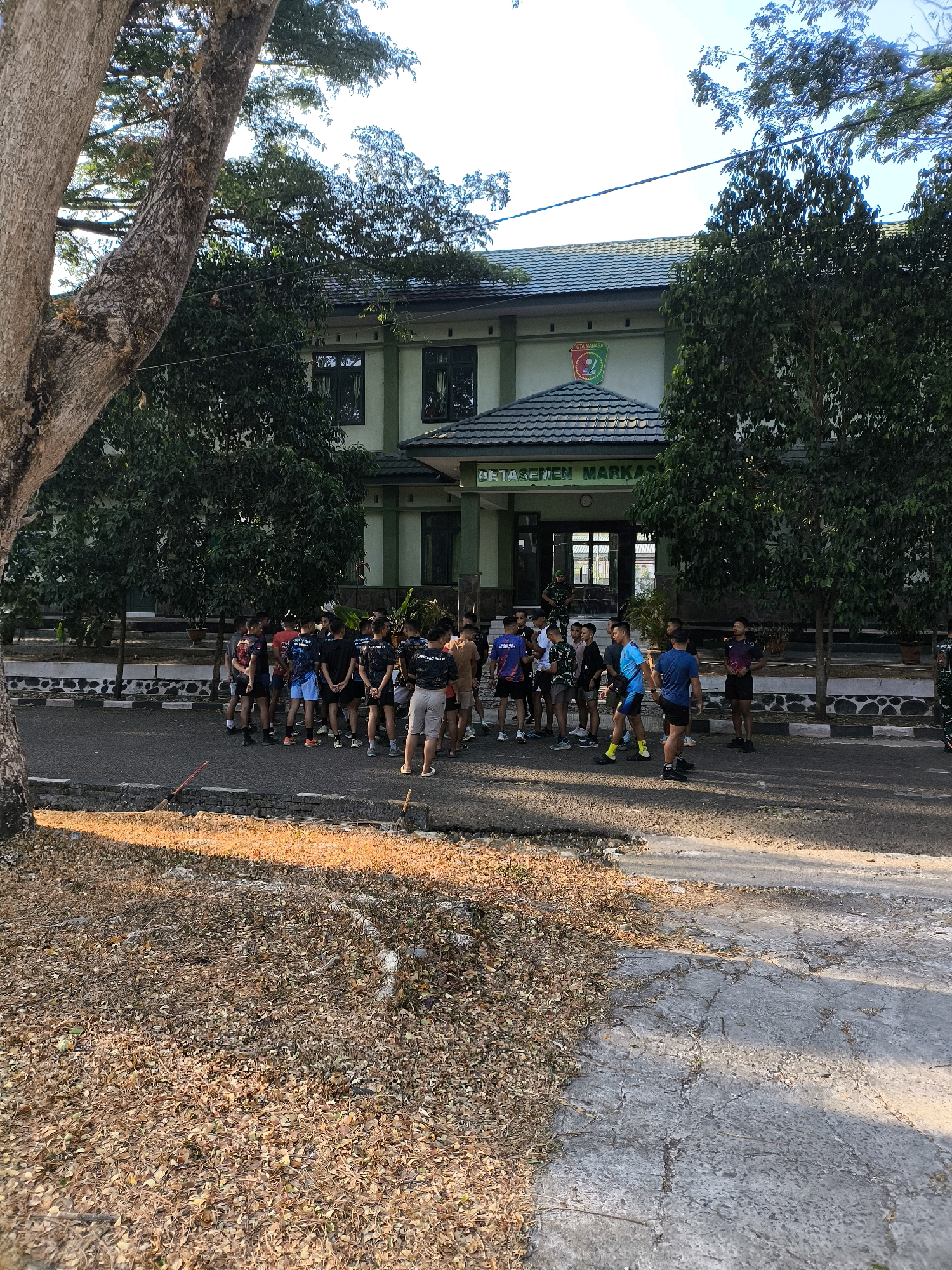 The image shows a group of approximately 25-30 young men, appearing to be in their late teens to early twenties, gathered on an asphalt road in front of a two-story building. They are casually dressed in athletic wear, including t-shirts, shorts, and sneakers. The men are standing in small groups, some facing each other in conversation, while others are looking towards the building. The building has a light green facade with dark green trim and a dark roof. A circular emblem with multiple colors is visible on the building's facade above the entrance. Text above the emblem appears to read "DETASEMEN MARKA". The environment is outdoors, with trees and foliage surrounding the building and the road. The ground in the foreground is covered with dry leaves and twigs. The lighting suggests it is daytime, with shadows cast by the trees and building.