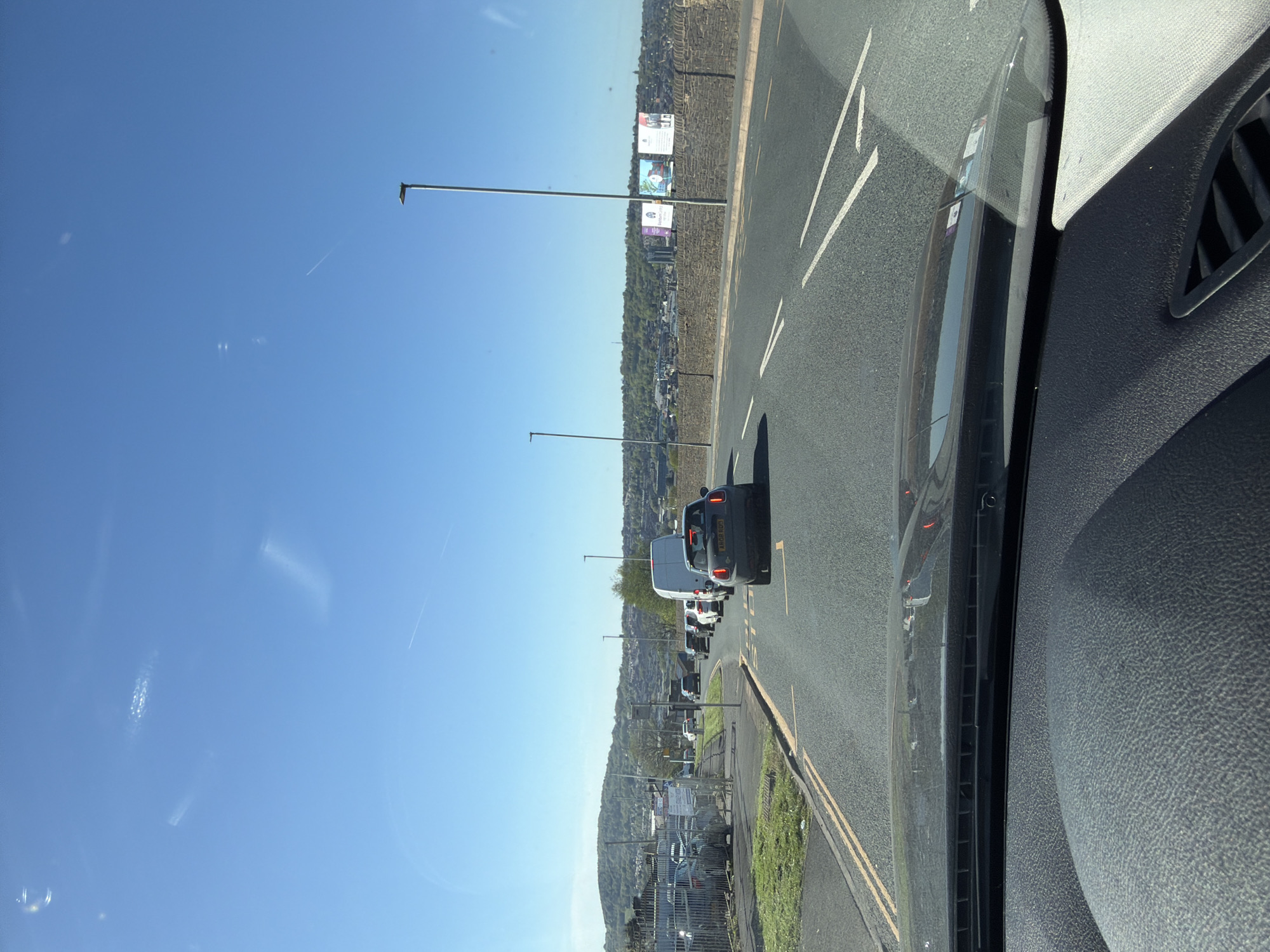 The image captures a scene from inside a vehicle, looking out through the front windshield onto a road in Blackburn, United Kingdom. The perspective is slightly angled, showing part of the vehicle's dark-textured dashboard and an air vent on the right.
Ahead, the road is visible with white dashed lane markings and double yellow lines along the curb. Several cars and a white van are in traffic, apparently stopped or moving slowly. The rear of a black car with the license plate 'AK08 RGO' is prominent in the mid-ground.
Beyond the road, a fence separates the road from a lower-lying area with parked cars. Further in the distance, a hillside rises, densely covered with trees and scattered buildings. Overhead, streetlights stand tall. On the hillside, several billboards are visible. One clearly displays "BLACKBURN College" with associated branding. Another billboard appears to advertise educational or vocational opportunities, possibly featuring text like "Future".
The sky is a clear, vibrant blue, indicating a bright, sunny daytime. A faint contrail from an aircraft is visible high in the sky. The overall impression is of a typical sunny day with urban traffic and a view extending to a green, built-up landscape.