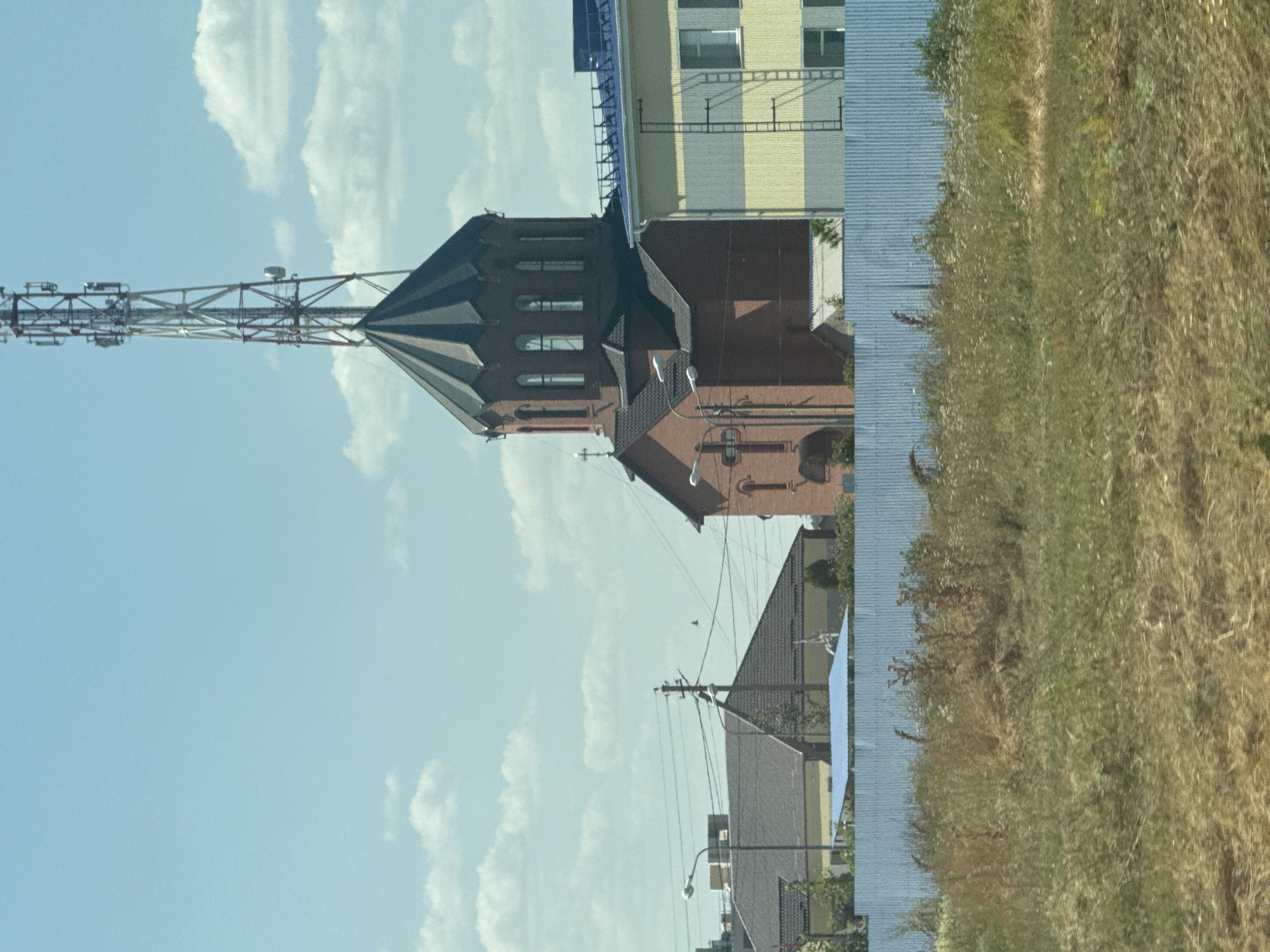 In the city of Shaumyanovsky, Russia, a cluster of buildings is visible under a bright, cloudy sky. The most prominent structure is a red brick church with a dark, multi-faceted dome topped with a cross. To the right of the church is a modern building with horizontal yellow and gray siding, featuring a visible external ladder and windows. A tall telecommunications tower stands behind and to the right of the church dome, with its metallic framework stark against the sky. In the middle ground, a blue-fenced area separates the buildings from a foreground of dry, tall grass and weeds. Other structures, including houses with dark roofs and power lines, are visible in the background.
