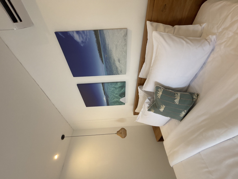This image depicts a modern bedroom in Canggu, Indonesia. The central subject is a neatly made bed with a light brown wooden headboard, featuring white sheets, a white duvet, four white standard pillows, and a single decorative green pillow with light-colored embroidered patterns. Above the headboard, two large vertical rectangular canvas prints are mounted on the white wall, showcasing beach scenes with clear turquoise ocean water, sandy shores, and distant trees under blue skies. The left print includes a visible white surfboard partially submerged in the water. A white wall-mounted air conditioning unit is positioned in the upper right, above the right print. In the upper left, a brown woven basket-style pendant light fixture hangs from the white ceiling, near a recessed ceiling spotlight. A light brown wooden bedside table is partially visible to the left of the bed. No individuals are present in the scene.