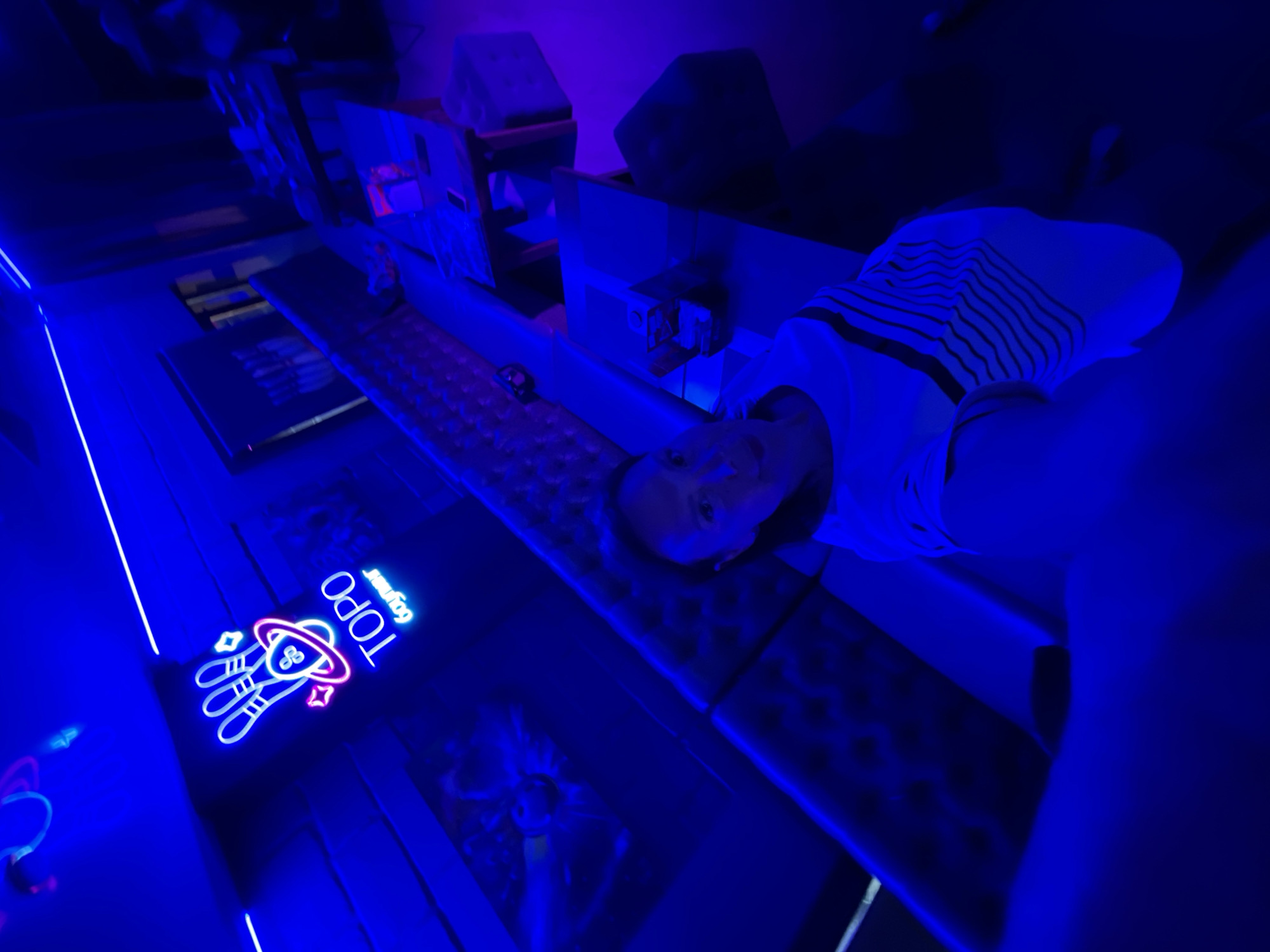 A person is lying on a black massage bed in a darkened room with blue lighting. The person is wearing a white shirt with black stripes. A neon sign that says "TOPO GYM" with a hand-and-UFO design is visible in the background. There are other pieces of equipment in the background, including a wooden structure with cushions. The location is Sochi, Russia.