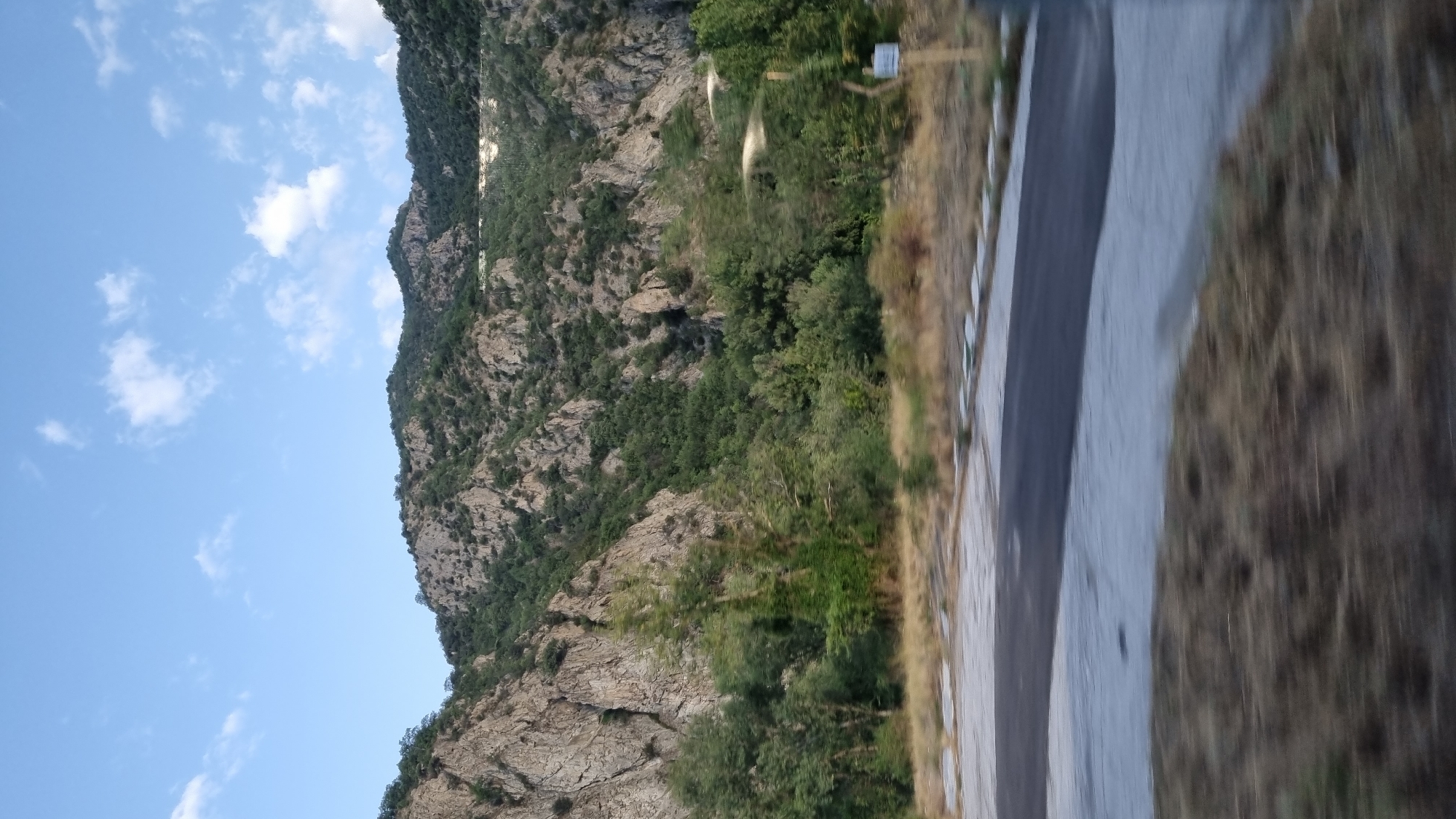 The image depicts a mountainous landscape with a road in the foreground. The mountains are rugged and covered with green vegetation, primarily trees and shrubs. The sky is blue with scattered white clouds. The road appears to be paved and has a light-colored border. There is dry grass and some smaller plants along the side of the road. A thin, dark pole with a white sign is visible on the right side of the image. The image appears to have been taken from a moving vehicle, as there is motion blur in the foreground, particularly in the dry grass. The overall impression is of a natural, somewhat wild environment, potentially on the outskirts of a town or city. Based on the context provided, this scene is located in Kresna, Bulgaria.