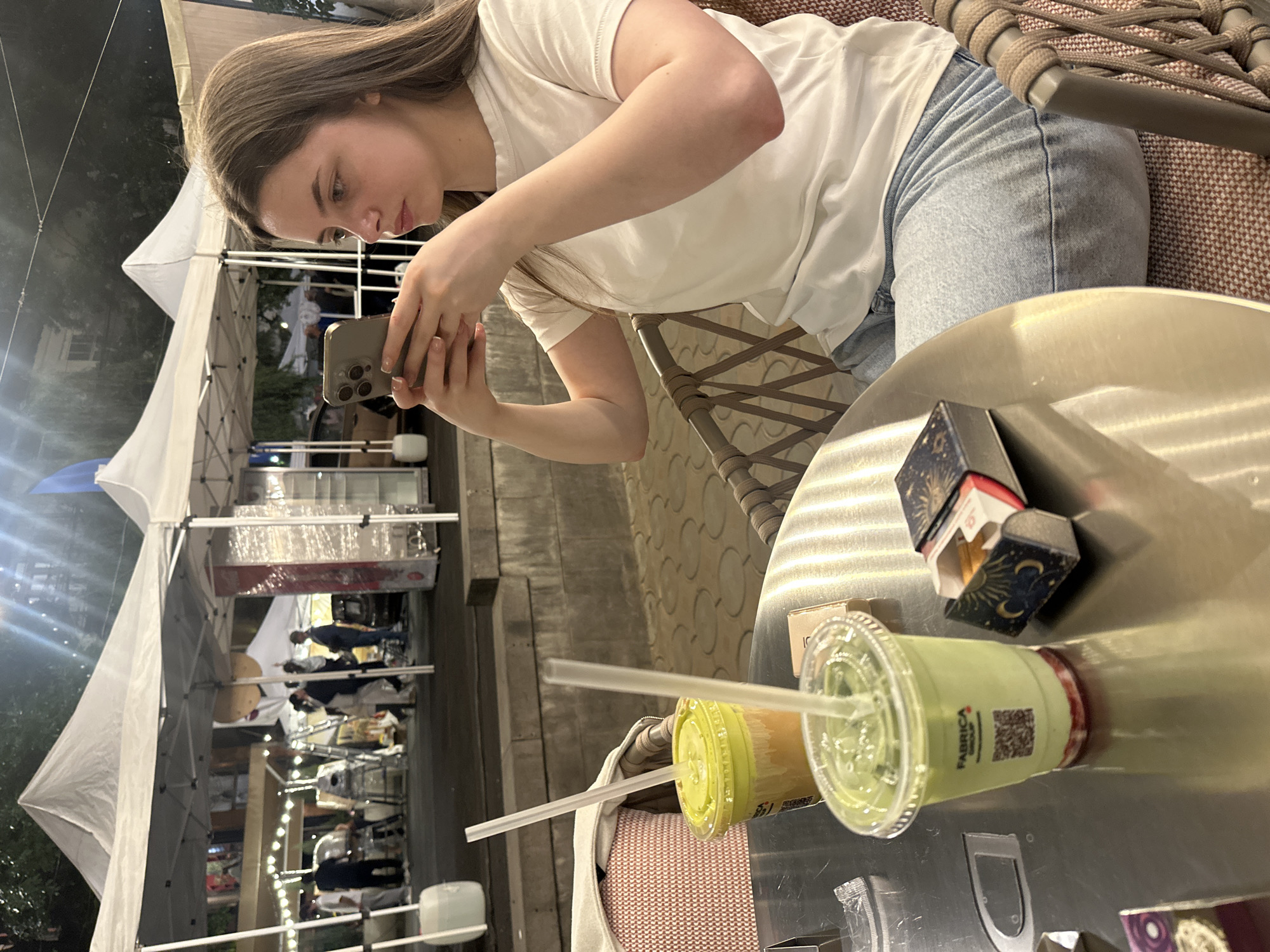 The image captures a young woman, appearing to be in her early twenties, seated at a metal table in an outdoor or semi-outdoor evening setting. She has long, light brown hair and is wearing a white short-sleeved t-shirt and light blue jeans. She is actively engaged in taking a photograph or video with a silver smartphone held in both hands, pointing it down towards the table. Her gaze is focused on the phone's screen.
On the table in front of her are two plastic cups with straws; one contains a light green beverage and the other a yellowish-orange drink. The green cup prominently displays the text "FABRICA" and a QR code. Nearby, there are two small boxes; one is dark blue or black with gold celestial designs (moons and stars) and features a small white label on top with "PABRICA" printed on it. The woman is seated on a chair with a woven backrest.
The background reveals a bustling outdoor market or event, characterized by multiple white canopy tents illuminated by strung lights. Silhouettes of other people are visible beneath these tents, suggesting vendor stalls or food stands. The time of day is clearly evening or night, indicated by the artificial lighting and the dark sky. The scene conveys a casual and relaxed atmosphere, likely occurring in Yerevan, Armenia, as indicated by the provided context.
