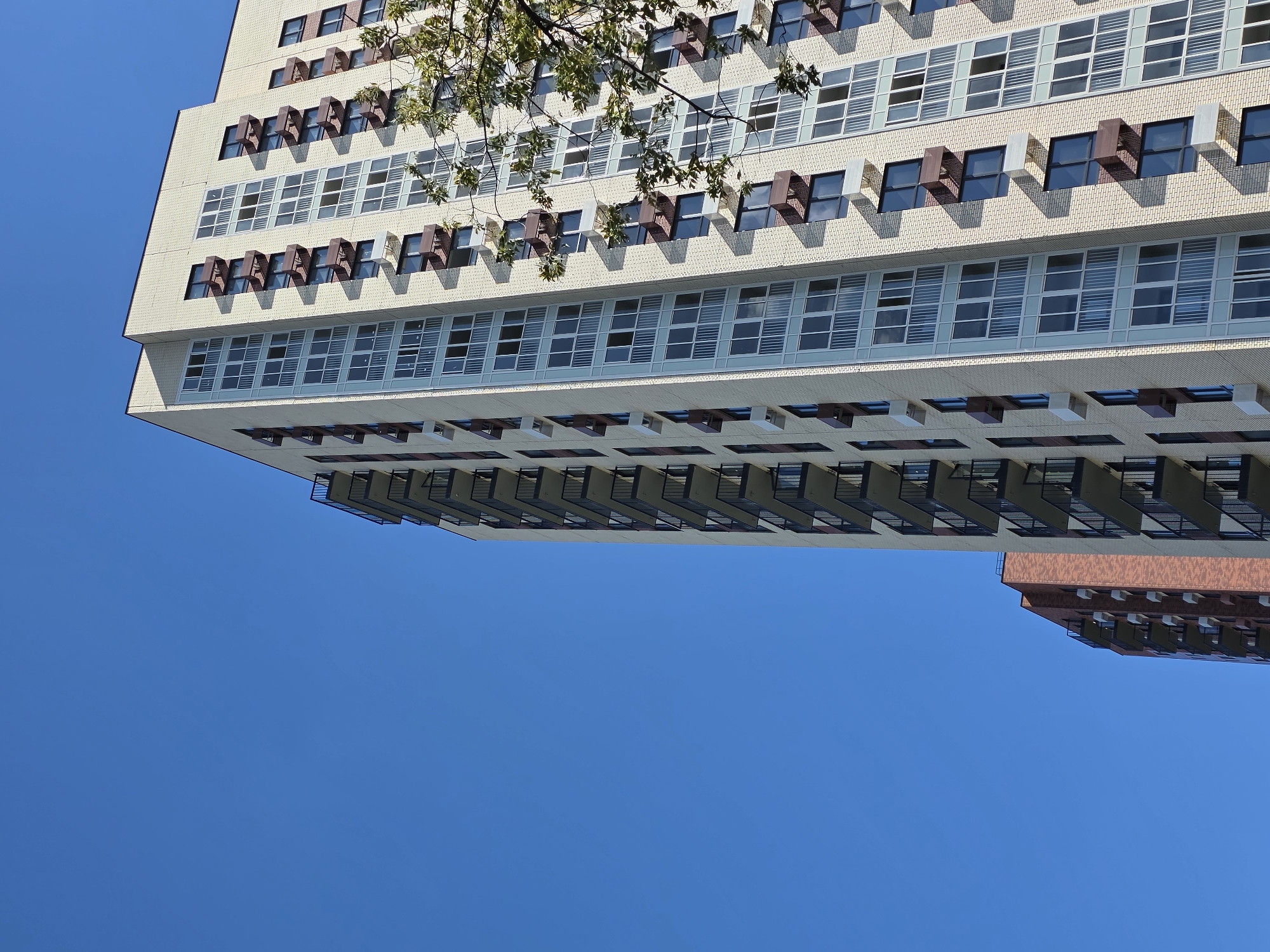 An upward-angle, medium shot captures a brightly lit, modern apartment building under a clear blue sky. The building's facade is primarily light-colored brick, with numerous large windows and balconies. Some balconies have dark metal railings, while others appear to be enclosed. Brown and white rectangular units, likely air conditioning units, are mounted below many of the windows. A second, taller building with a reddish-brown brick facade is visible behind and to the left of the main building, also featuring balconies. A tree branch with green leaves partially obscures the upper right corner of the main building. The image appears to have been taken in Moscow, Russia, given the architectural style. No people are visible in the frame.