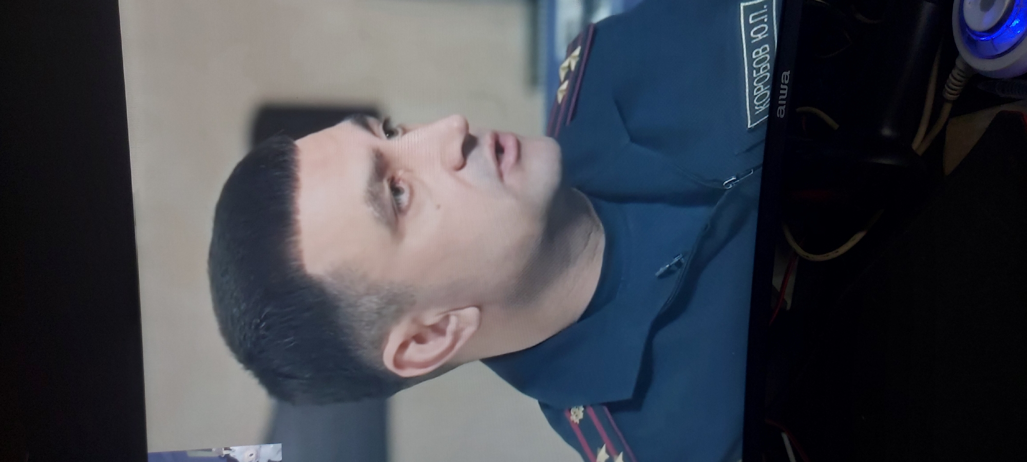 A man with a short, dark haircut is facing towards the right. He is wearing a dark blue uniform with a name tag in Cyrillic script that reads "КОРОБОВ Ю.П.". The background behind him is blurred and indistinct. To the right of the man, out of focus, are electronic devices and cables, suggesting an indoor setting.