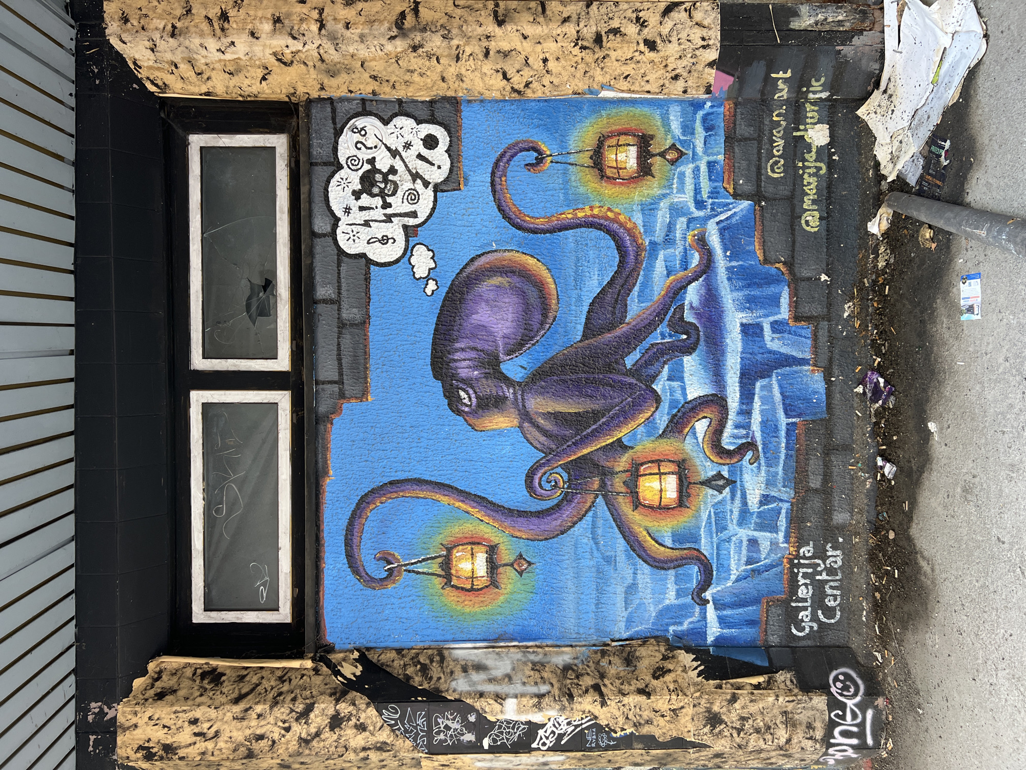 A piece of street art is painted on a wall on the streets of Belgrade, Serbia. The art depicts a purple octopus on a blue background, holding a lantern in two of its tentacles. Above the octopus is a white thought bubble that contains a skull and crossbones, question marks, and exclamation points. The name "Galerija Centar" is written in white on the bottom right corner. Next to the writing is a light gray sidewalk littered with small debris. To the right of the artwork, two social media usernames "ava.n.art" and "@marija.djuric" are written in yellow. The wall is next to a black-framed window with two panes, both of which are broken. Next to the window is a black tiled wall and white siding.