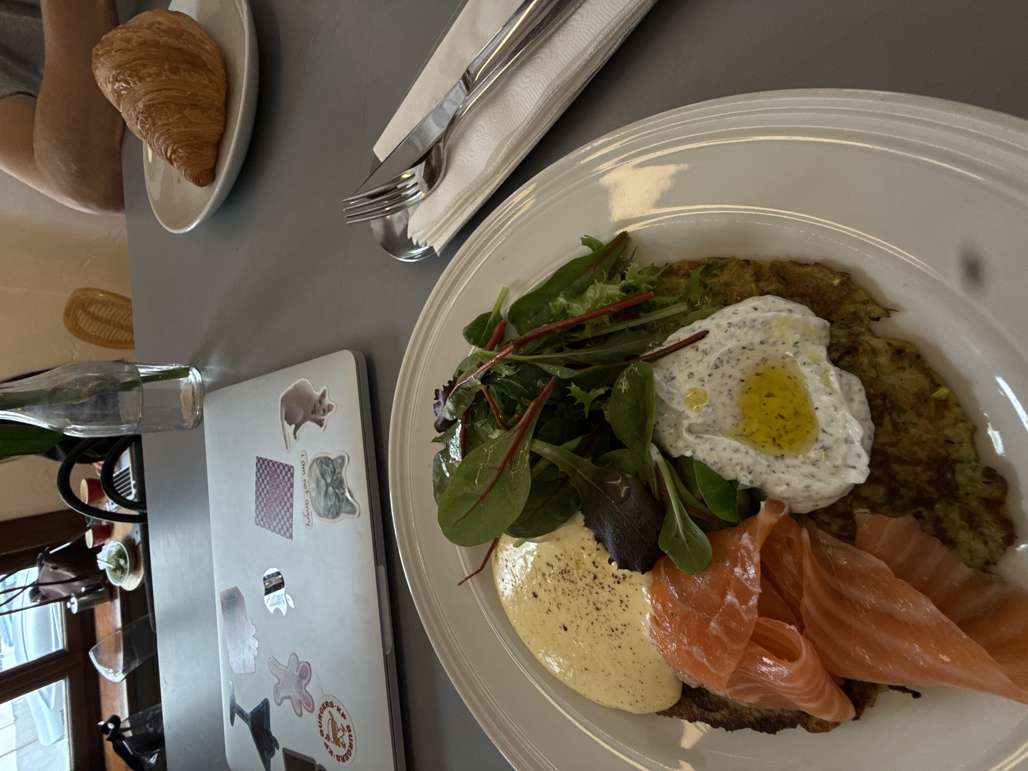 A top-down shot displays a meal served on a white plate. The meal consists of a latke topped with greens, a white sauce (possibly hollandaise), and slices of salmon. Nearby, there is a croissant on a separate plate, along with silverware wrapped in a napkin. A laptop with stickers is also visible, alongside a vase with plant stems. The scene is probably set in a restaurant or cafe in Belgrade, Serbia.