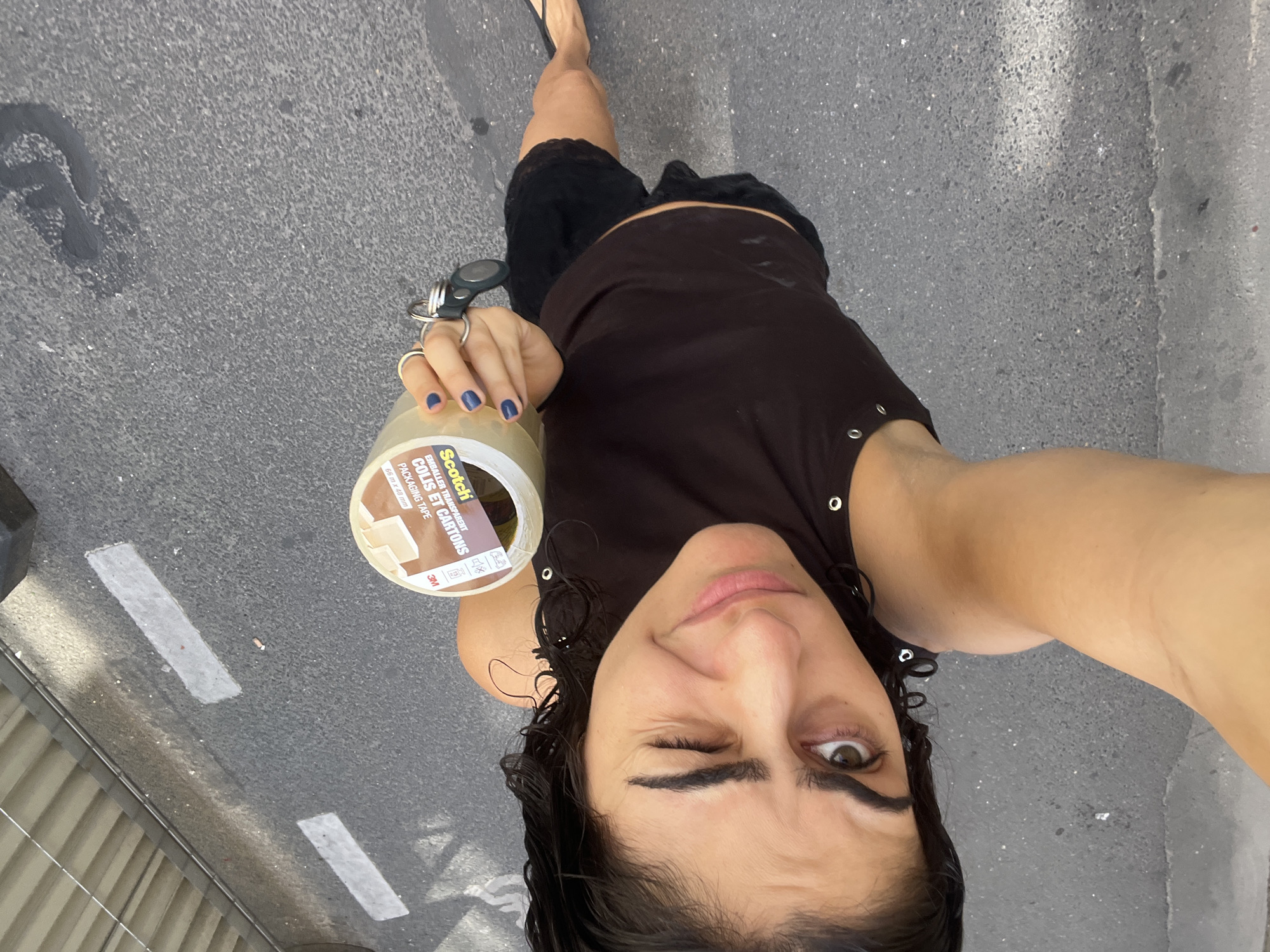 In a tilted, close-up selfie taken outdoors on what appears to be a street in Paris, France, a person is holding a roll of Scotch packaging tape. The tape is transparent and has visible text, including "EMBALLER TRANSPARENT COLIS ET CARTONS PACKAGING TAPE" and "3M". The person's right hand, adorned with blue nail polish and rings, is gripping the tape. They are wearing a dark tank top and black lace-trimmed shorts. Their facial expression is a playful wink, with their right eye open and their left eye closed, and a slight smile. The background shows a grey asphalt surface with some white markings, suggesting a road or sidewalk. The lighting indicates it is daytime, likely sunny given the brightness and sharp shadows, though no clear weather information is available. The overall impression is a candid, lighthearted moment captured during an activity involving packaging or moving.