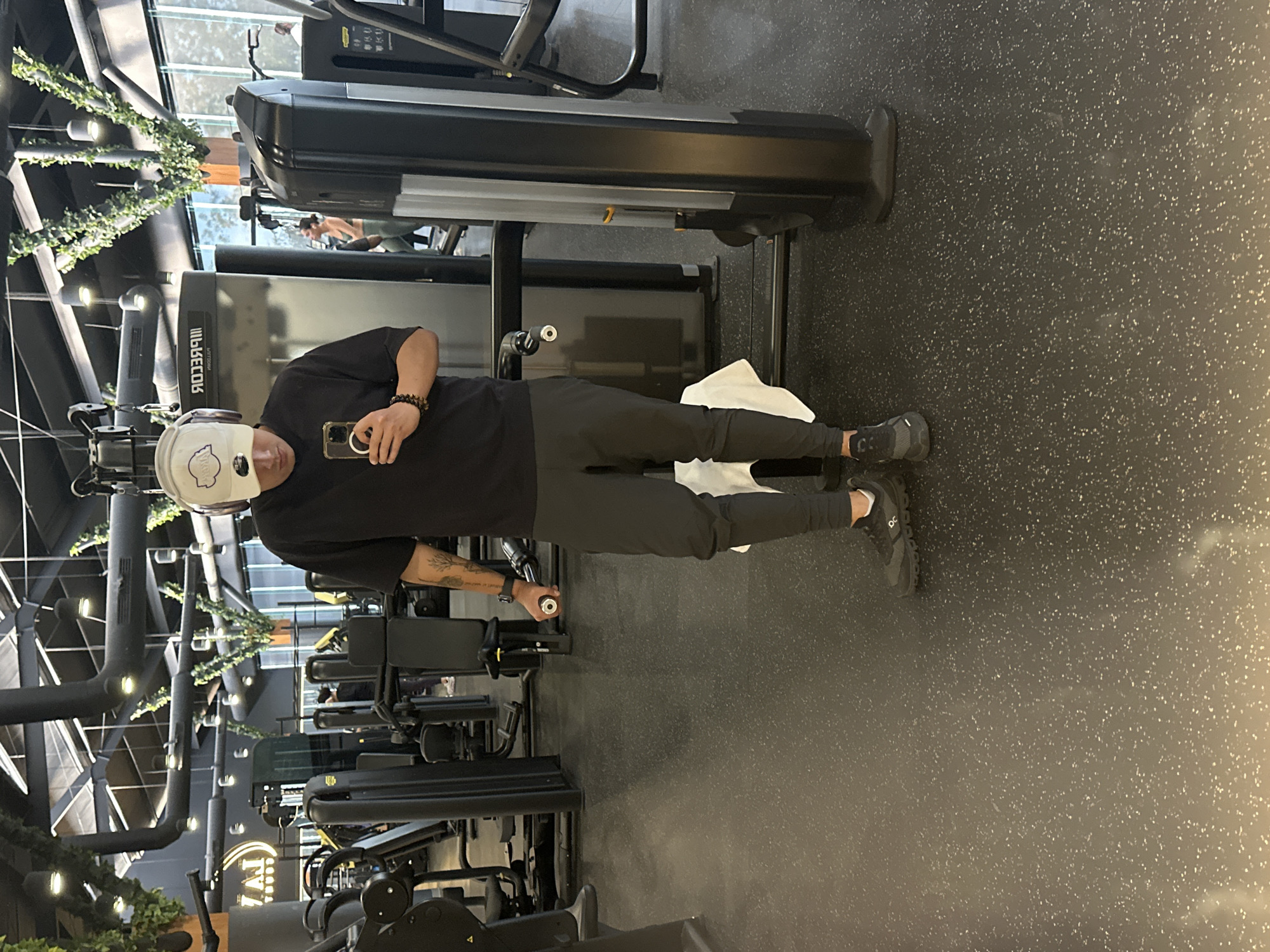 A person wearing a white hat and a black t-shirt stands in a gym. They are holding a cell phone in their right hand and grasping a weight machine handle with their left hand. They are wearing dark athletic pants and dark sneakers. Behind them is a weight machine, a treadmill, and other gym equipment. The gym has black flooring. The location appears to be indoors and well-lit. There is no evidence to support the location being San Bernardino Tlaxcalancingo, Mexico.