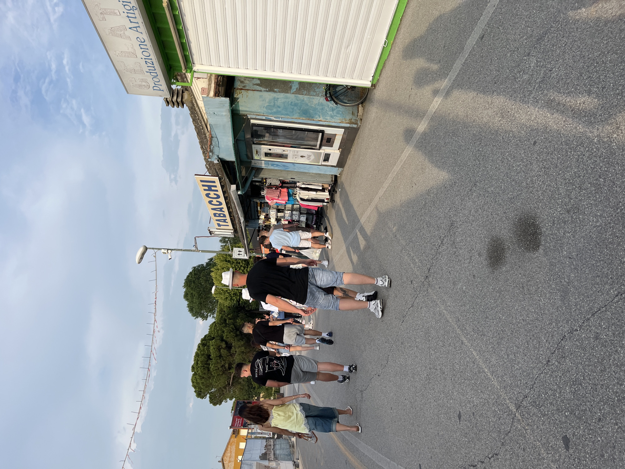 The image shows a street scene in Punta Sabbioni, Italy. A line of people walks along the edge of a street beside a small shop with a "TABACCHI" sign and a "Produzione Artigianale" sign above. The shop has a closed white roller shutter and vending machines on the side. People are dressed in casual summer clothing. The sky is visible above trees and a light pole with decorative lights. The asphalt road is cracked in places.