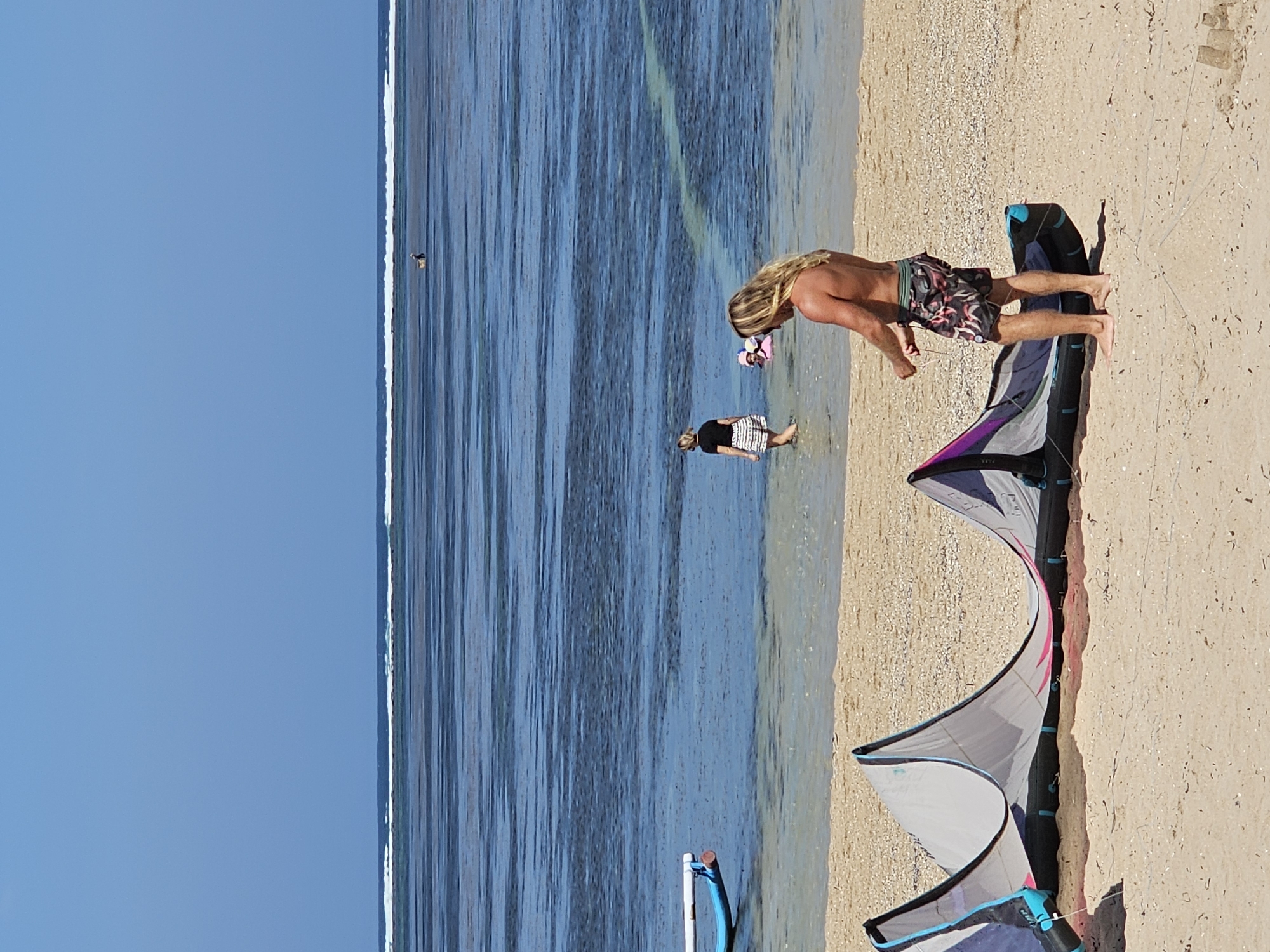 A man with long blonde hair, wearing patterned swim shorts, stands on a sandy beach in Denpasar, Indonesia, facing away from the camera and towards the ocean. He is holding a kite line and appears to be preparing to launch a kite, which is partially visible on the sand in the foreground. The kite is large and multi-colored, with shades of gray, purple, and pink. In the shallow, clear water, a woman in a black top and striped shorts wades, and further out, a person in a pink float is visible. The ocean stretches to the horizon under a clear blue sky, with a distinct line of breaking waves in the distance. A blue and white boat is moored in the water to the left of the frame. The beach is sandy and sparsely populated.