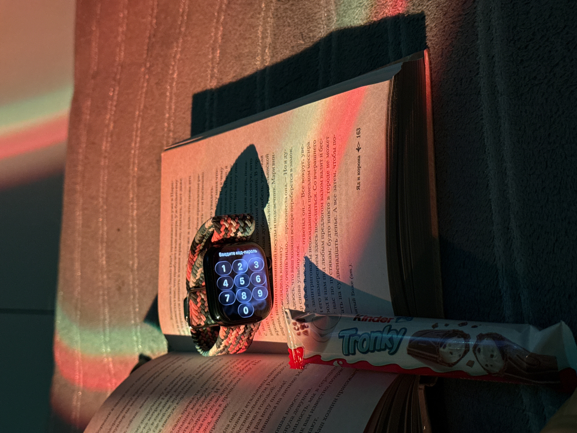 The image shows an open book with an Apple Watch resting on the left side and a Kinder Tronky chocolate bar on the bottom right. The watch display shows a passcode entry screen. The book's pages are filled with Russian text. The scene is illuminated by warm, colorful light. The background appears to be a textured surface.