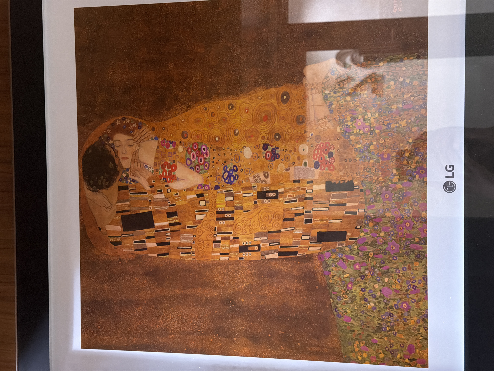 The image shows a reproduction of Gustav Klimt's "The Kiss". The painting depicts a couple embracing, their bodies intertwined in elaborate robes. The man has dark hair and is kneeling to kiss the woman. The woman has red hair adorned with flowers and is resting her hands on his. Their figures are set against a gold background. The bottom right corner of the image is an LG television set.
