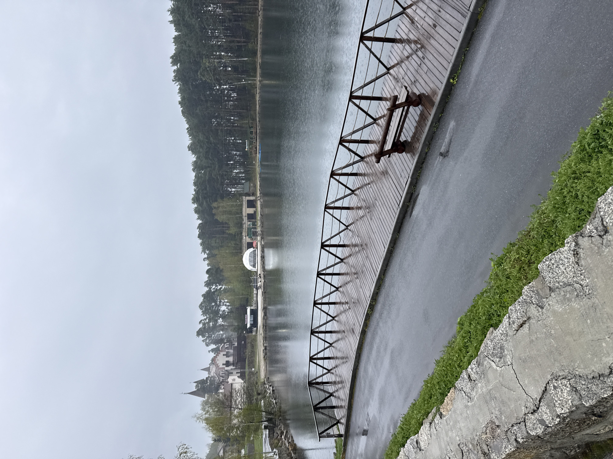 A photograph shows a lakefront scene in Turgoyak, Russia, on a cloudy or rainy day. A wooden boardwalk with a metal railing runs along the edge of the water. On the boardwalk, there is a simple wooden bench. To the right of the boardwalk, a wet asphalt path is visible, bordered by grass and a low, cracked stone wall. Across the lake, trees line the shore, behind which are buildings including a white dome-like structure, darker buildings, and a building with multiple spires. The overall atmosphere is overcast.