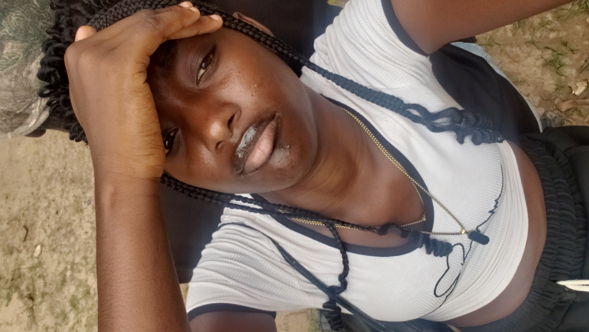 A young Black woman with braided hair is taking a selfie outdoors. She is wearing a white crop top with black trim and a gold chain necklace with a dark pendant. Her right hand is raised to her forehead, and she is looking towards the camera with a neutral expression. The background appears to be an outdoor setting with dirt and sparse vegetation, consistent with a location in the city of Kaduna, Nigeria.