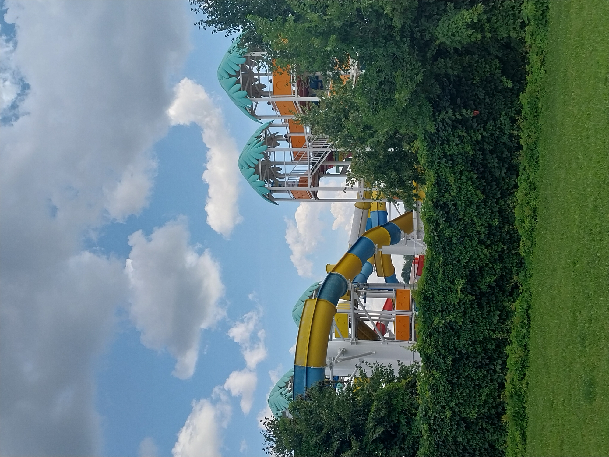 The image depicts a section of the "Happy Day" water park in Dnipro, Ukraine, on a partly cloudy day. In the foreground, a well-maintained lawn transitions into a dense, dark green hedge. Behind the hedge, the lower portions of brightly colored water slides are visible, with segments of yellow and blue tubes curving into the scene. Further back, a white structure supports multiple levels of the water park, adorned with tropical-themed canopies resembling palm fronds. Orange and red sections are also visible, suggesting further slide elements or splash zones. Several individuals are discernible on the elevated platforms of the water park, appearing to be waiting or observing. The sky above is a blend of bright blue with scattered white and gray clouds. The overall scene captures a recreational facility during operational hours.