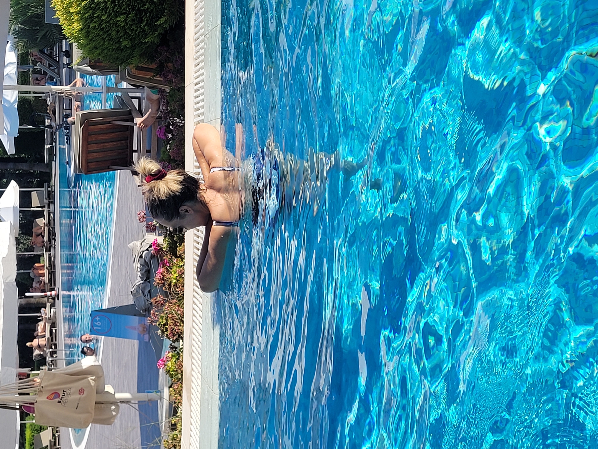 A woman with blonde hair pulled into a bun, wearing a blue and white patterned bikini, rests her arms on the edge of a swimming pool. Her back is to the camera as she gazes towards the far end of the pool. The pool water is a vibrant turquoise, reflecting the bright sunlight. In the background, several lounge chairs are arranged on a paved area beside the pool, some occupied by sunbathers. Lush green bushes and palm trees frame the scene. A white umbrella shades a section of the lounge area. Further in the distance, more pool areas and resort buildings are visible. A sign with "16+" is placed near the pool's edge. This scene is consistent with a resort in Manavgat, Turkey, known for its numerous hotels and recreational facilities along the Mediterranean coast.