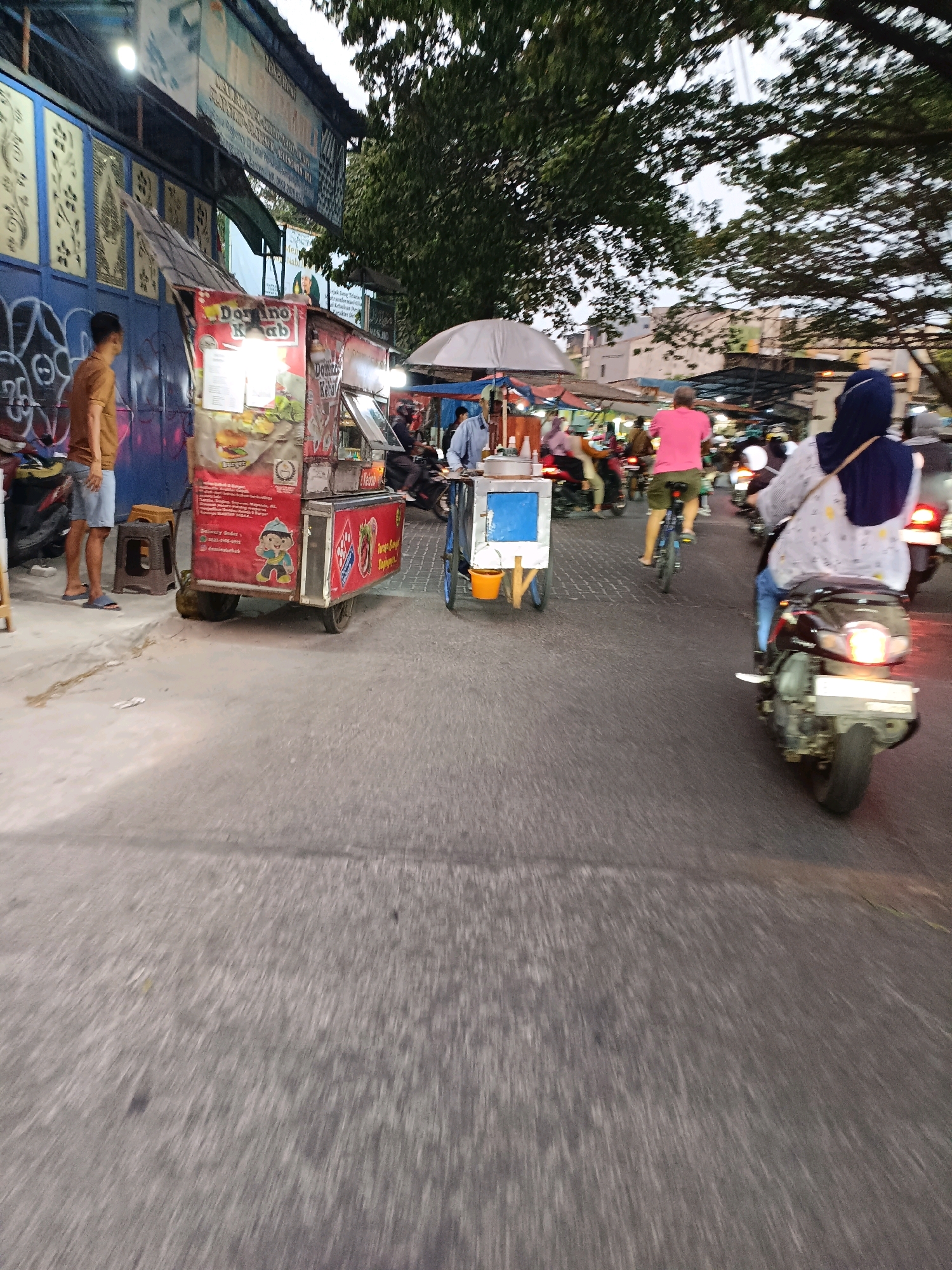 The image depicts a street scene in Gelam Jaya, Indonesia, with a focus on a food cart and surrounding activity. A person in a brown shirt and blue shorts stands near the food cart, which is red and white, displaying a menu with images of burgers. To the right, a person on a bicycle wearing a pink shirt is visible. Further to the right, a person wearing a blue hijab and a patterned shirt rides a motorcycle. The street is paved and appears to be a busy thoroughfare with other vehicles and a food cart in the background. Trees with dense foliage line the street, and the sky is overcast. The overall scene suggests a market or commercial area with street food vendors and a mix of pedestrian and vehicle traffic.