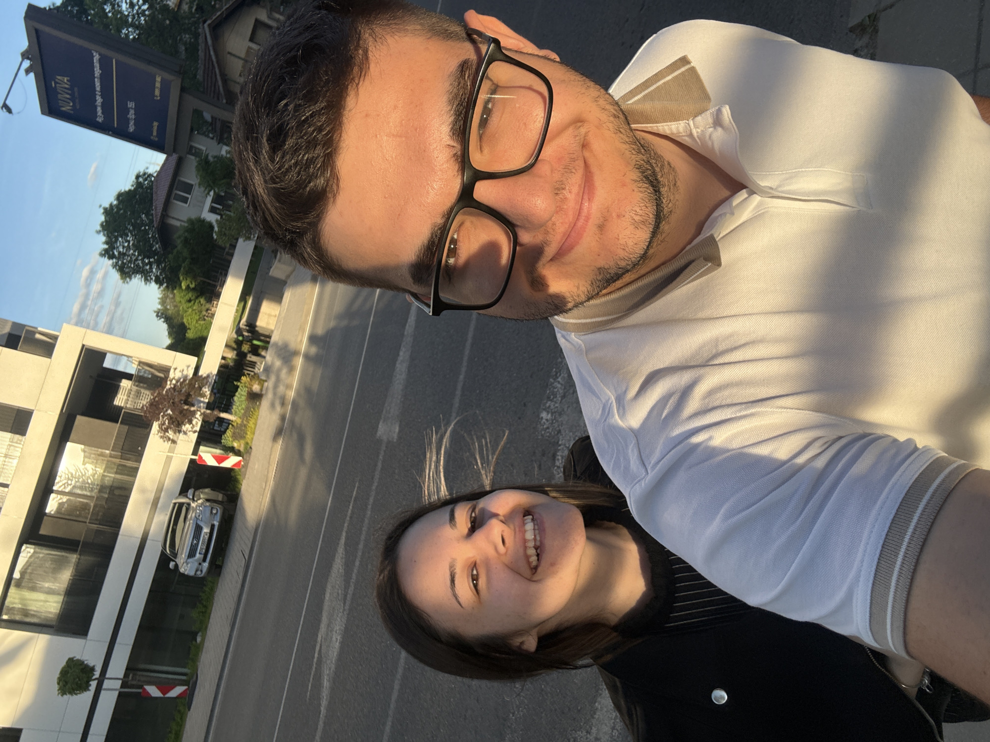 The image is a selfie of two people. The person holding the camera is a man wearing glasses and a white polo shirt. Next to him is a woman with dark hair. Both are smiling. The background shows a city street, and a sign says "NUVIVA". Based on the sign and the general architecture, the location is likely in Sofia, Bulgaria. A parked car is visible near a modern building. The time of day appears to be late afternoon or early evening, given the lighting.