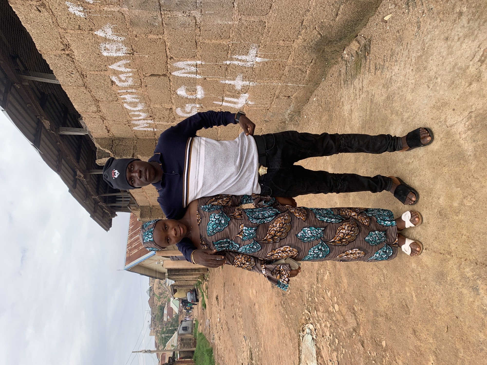 This image depicts two individuals, a male and a female, standing side-by-side on an unpaved, reddish-brown dirt road in the city of Jos, Nigeria.
The female, positioned on the left, wears a long, off-the-shoulder dress featuring a brown background with prominent green, teal, and orange leaf-like patterns. She wears a matching headscarf and white sandals. Her left hand holds a light-colored clutch or purse.
The male, positioned on the right, has his right arm draped over the female's left shoulder. He wears a navy blue long-sleeved polo shirt with a white and brown horizontal stripe across the chest, black ripped jeans, and black slide sandals. A black beanie with a red and black heart-like emblem is on his head. A gold-colored belt buckle with a Medusa head design is visible, along with a dark-colored watch on his left wrist. His left hand is seen tugging at the hem of his shirt.
To the right of the individuals is a rough, tan-colored concrete block wall. White spray paint graffiti on the wall reads "MUCHIGABA" at the top, followed by "GCM" and "4+4+4", with paint streaks running downwards. Above this wall, a structure with a corrugated metal roof, exposed wooden beams, and mesh fencing is visible.
To the left of the individuals, the unpaved dirt road extends into the background, flanked by various single-story residential buildings with corrugated metal roofs. In the far background, a rocky, hilly terrain dotted with more residential structures is visible under an overcast sky.