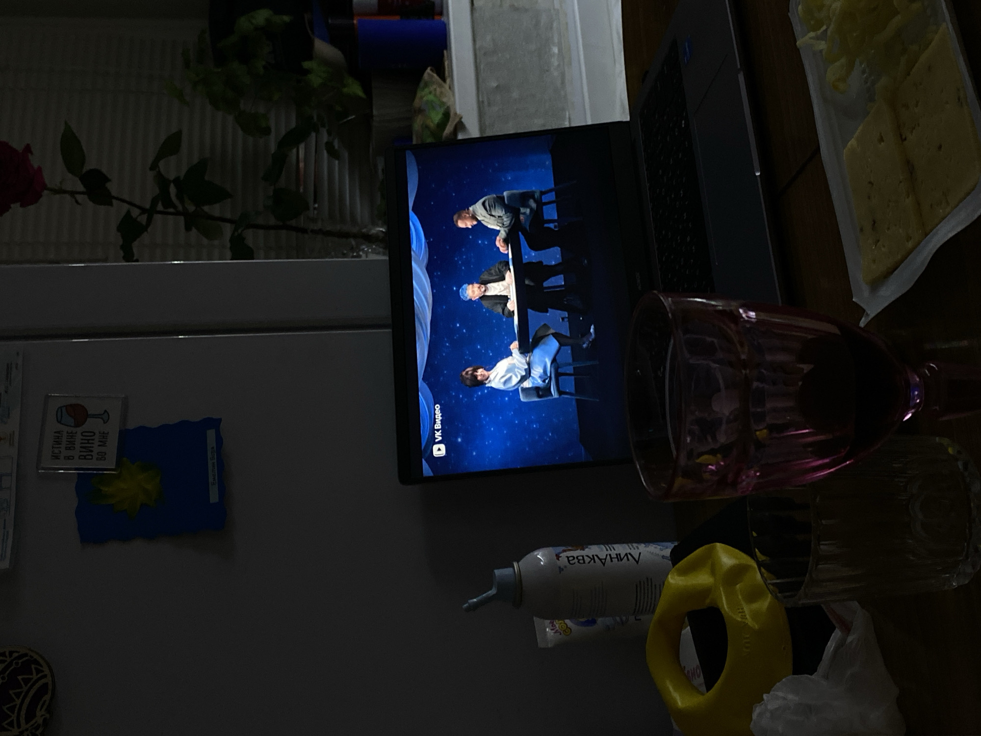 A dimly lit indoor scene shows a laptop screen displaying a video, likely in Russian, featuring three people on stage against a blue backdrop. The scene is cluttered with objects including a plate of cheese, drinking glasses, and a bottle of what appears to be cosmetic product. There is also a yellow handle object near the bottle. In the background, visible to the left, is a refrigerator with magnets attached. The location in Votkinsk, Russia cannot be directly confirmed based on the image alone, but the Russian text suggests it as a possibility.