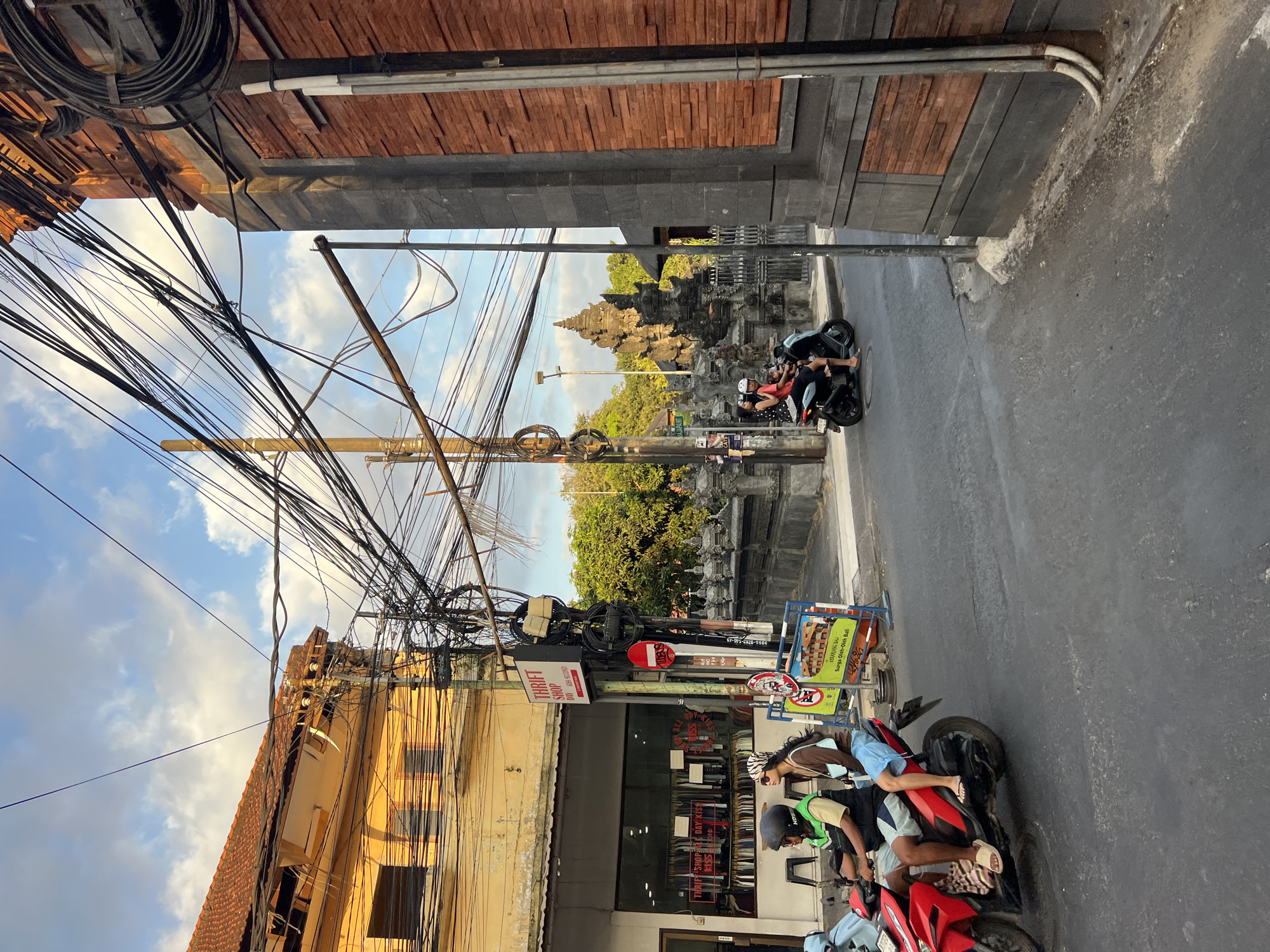 The image depicts a street scene in Kuta, Indonesia, characterized by a dense network of overhead electrical wires crisscrossing the sky. On the left, a yellow building with a tiled roof houses a thrift shop with its interior visible, displaying racks of clothing. A red motorcycle with two occupants, a driver wearing a helmet and a passenger with sunglasses, travels along the asphalt road. In the middle ground, a stone temple-like structure with intricate carvings is visible behind a low stone wall. Another motorcycle with two occupants is proceeding in the same direction on the right side of the road. The right side of the image is dominated by a building with a dark stone base and a brick facade. The sky is partly cloudy, with the sun casting a warm glow on the scene.