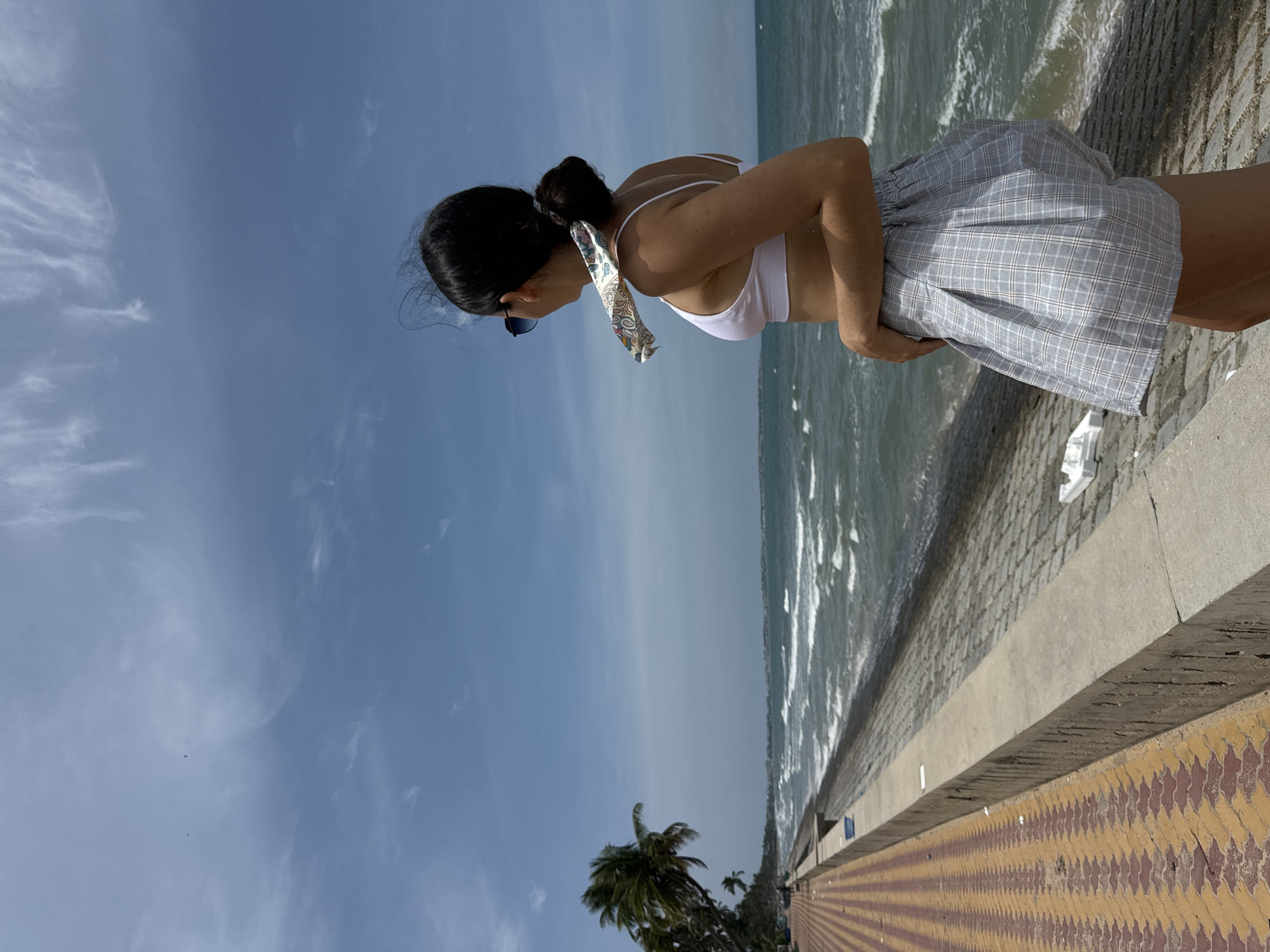 A woman stands with her back to the camera, facing the ocean. She is wearing a white bikini top and a grey and white plaid skirt. A patterned scarf is tied around her hair. She is standing on a concrete wall that separates a paved walkway from a rocky embankment leading to the sea. Palm trees are visible in the background, along with a distant coastline. The sky is blue with scattered white clouds. The scene appears to be a coastal area in Phu Thuy Ward, Vietnam, likely a promenade or seawall.
