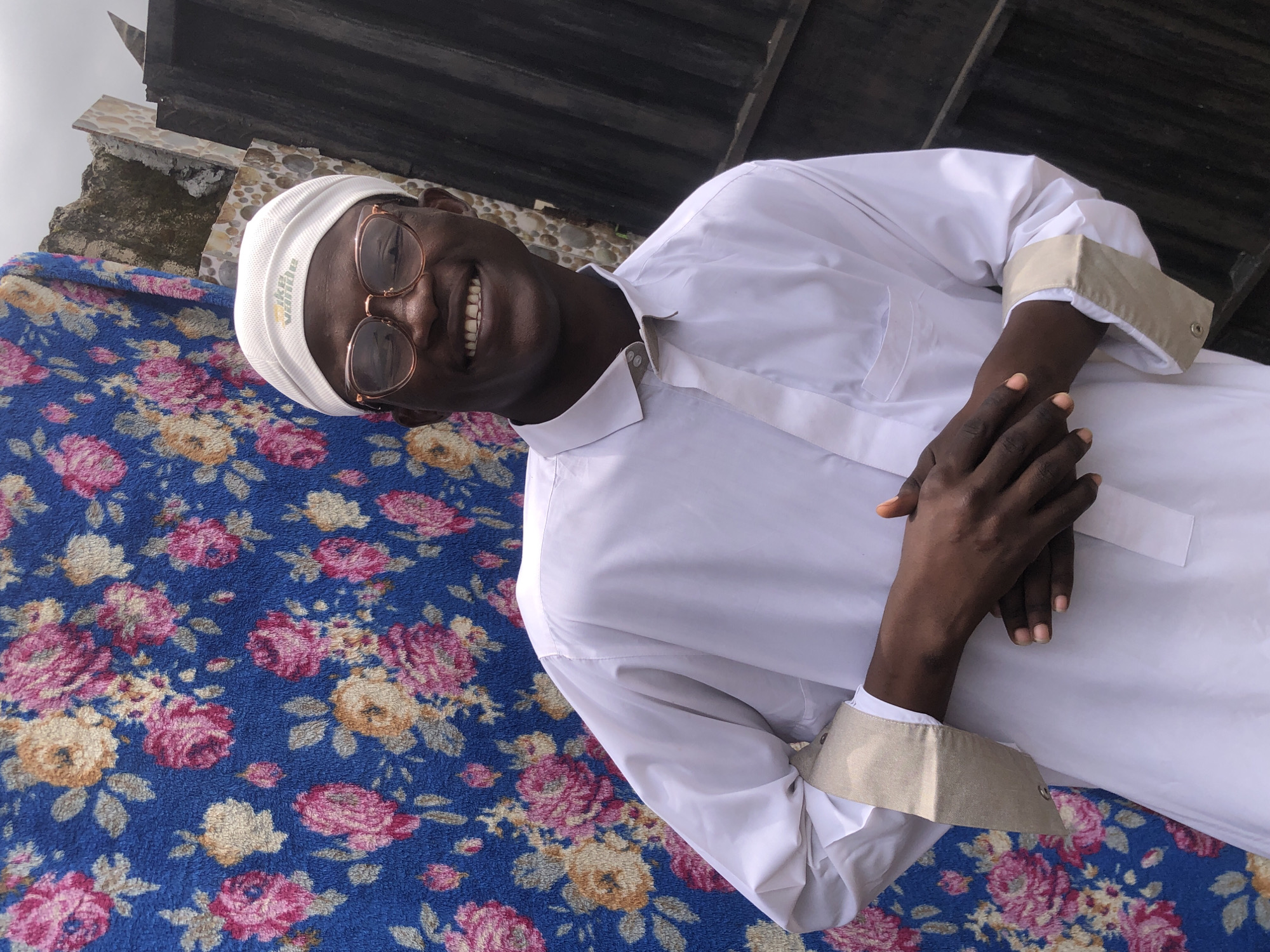 In Ado, Nigeria, a young Black man is captured in a medium shot, smiling warmly at the camera. He wears a white cap with a subtle logo and clear-rimmed glasses, highlighting his expressive eyes. His attire consists of a crisp white long-sleeved shirt with beige accents on the cuffs and collar. His hands are clasped in front of him, with one hand resting atop the other. Behind him, a vibrant blue backdrop adorned with a floral pattern of pink, yellow, and beige flowers creates a striking contrast. To the right, a dark, textured surface, possibly a fence or wall, adds a rustic element to the scene. The overall lighting suggests an outdoor setting with diffused natural light.