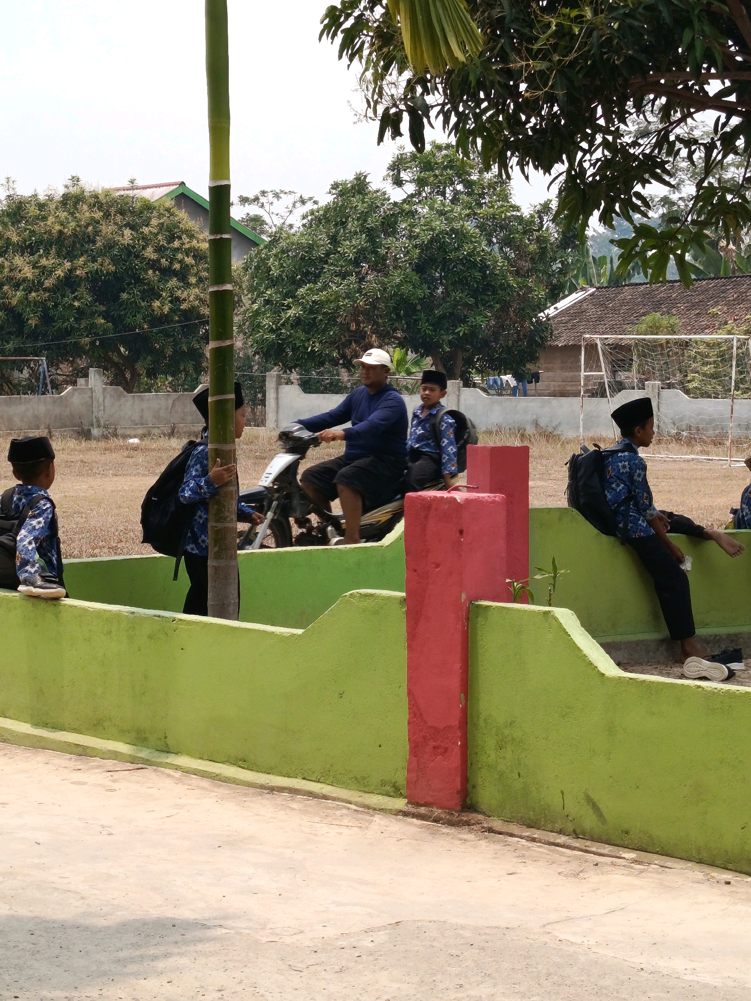 The image depicts an outdoor scene in what appears to be a schoolyard or similar communal area in Megang Sakti V, Indonesia. A man in a blue shirt and dark shorts, wearing a white cap, is riding a motorcycle. He is accompanied by three young boys, all wearing traditional Islamic skullcaps and patterned blue shirts. Two of the boys are seated on the back of the motorcycle, and one is standing beside it. Another boy, also wearing a skullcap and blue patterned shirt, is seated on a low, bright green wall. This wall is adorned with red pillars. In the background, a fence encloses a grassy field with a soccer goal. Trees with lush green foliage and houses with tiled roofs are visible behind the fence. A tall, green bamboo pole stands prominently in the foreground, next to the boy on the wall. The ground is a mixture of dry grass and a paved surface. The sky is overcast.