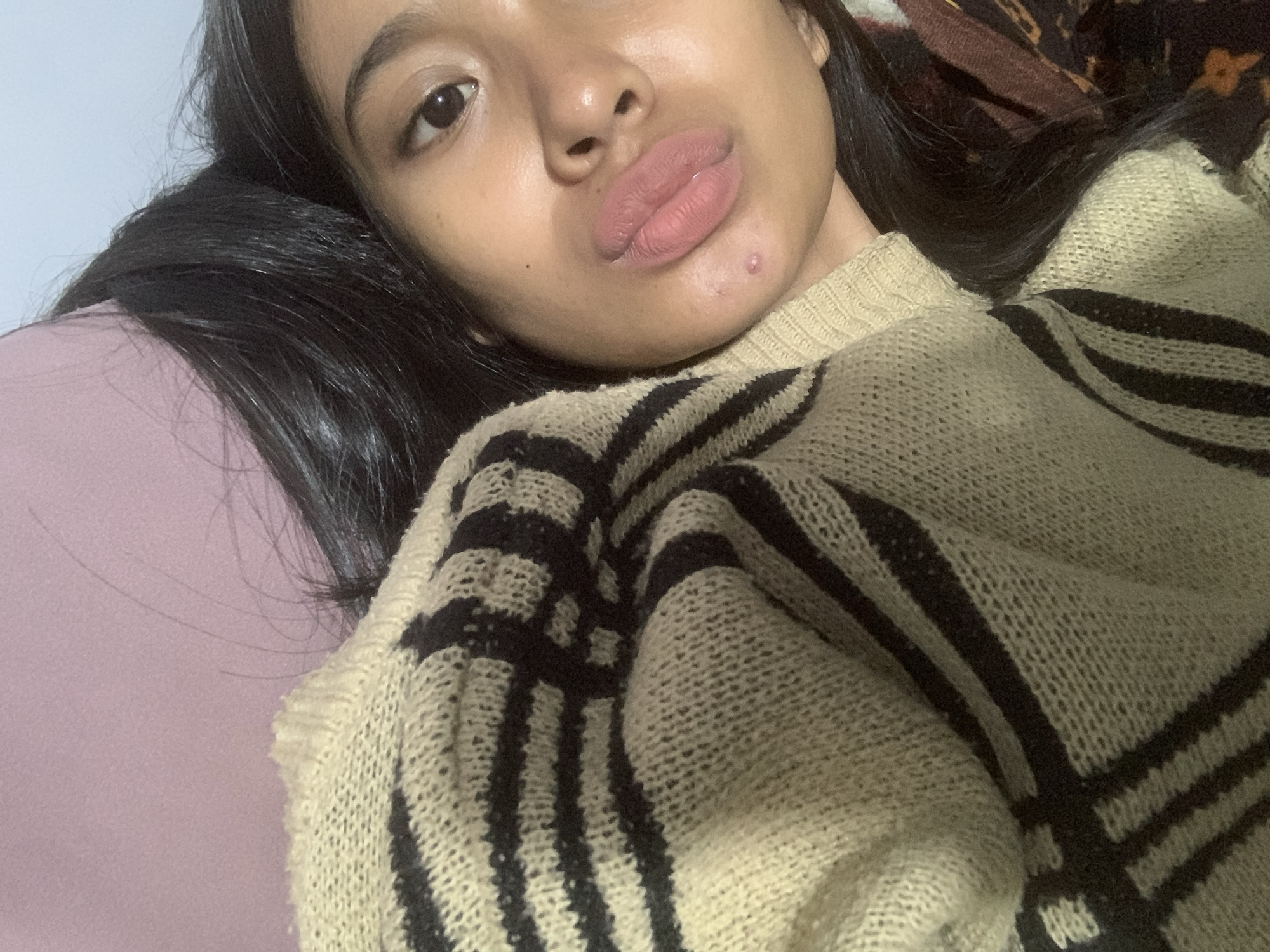A close-up selfie of a young woman with fair skin, brown eyes, and black hair, wearing a beige and black striped knit sweater. She has a small blemish near her lower lip. She is lying down, with her head resting on a light pink pillow. The background includes a glimpse of a dark brown patterned fabric. The location appears to be indoors, possibly in a residential setting in Sungai Meriam, Indonesia.